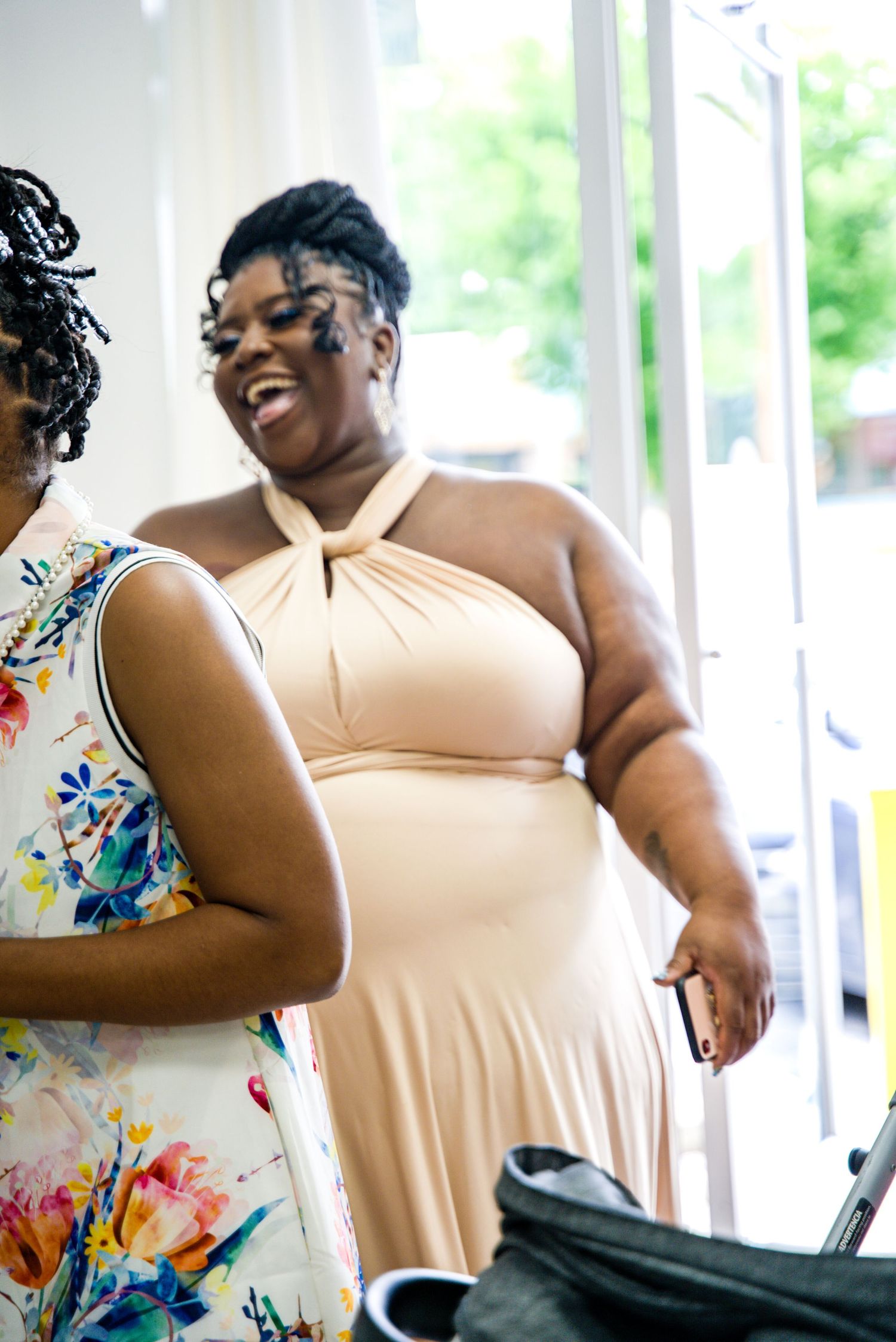 Laughing pregnant woman in beige halter dress chatting with friend at baby shower.