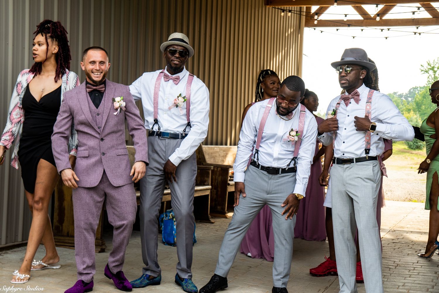 Groomsmen in gray pants and pink suspenders dancing joyfully at outdoor rustic barn wedding venue.