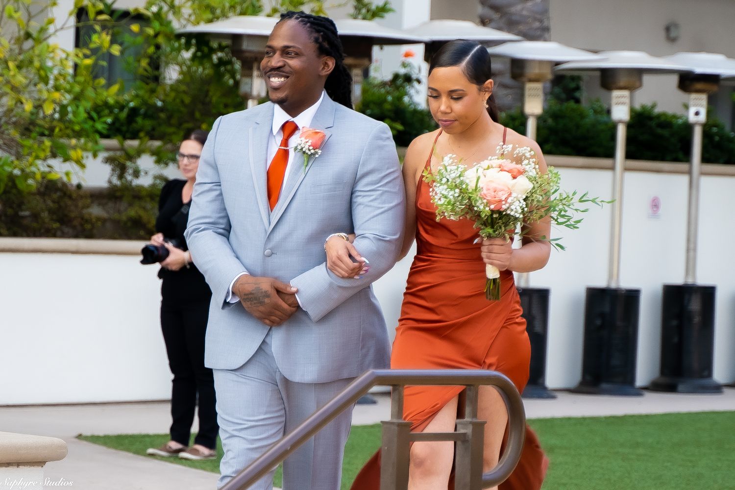 Groomsman in gray suit escorts bridesmaid in rust orange dress down wedding aisle.