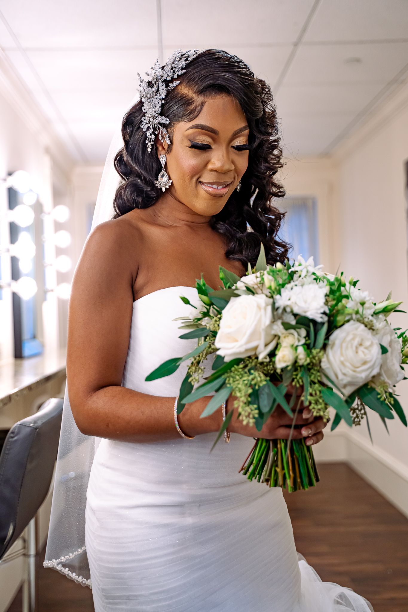 Radiant Black bride in strapless white gown holding white rose bouquet with greenery, adorned with crystal hair piece.