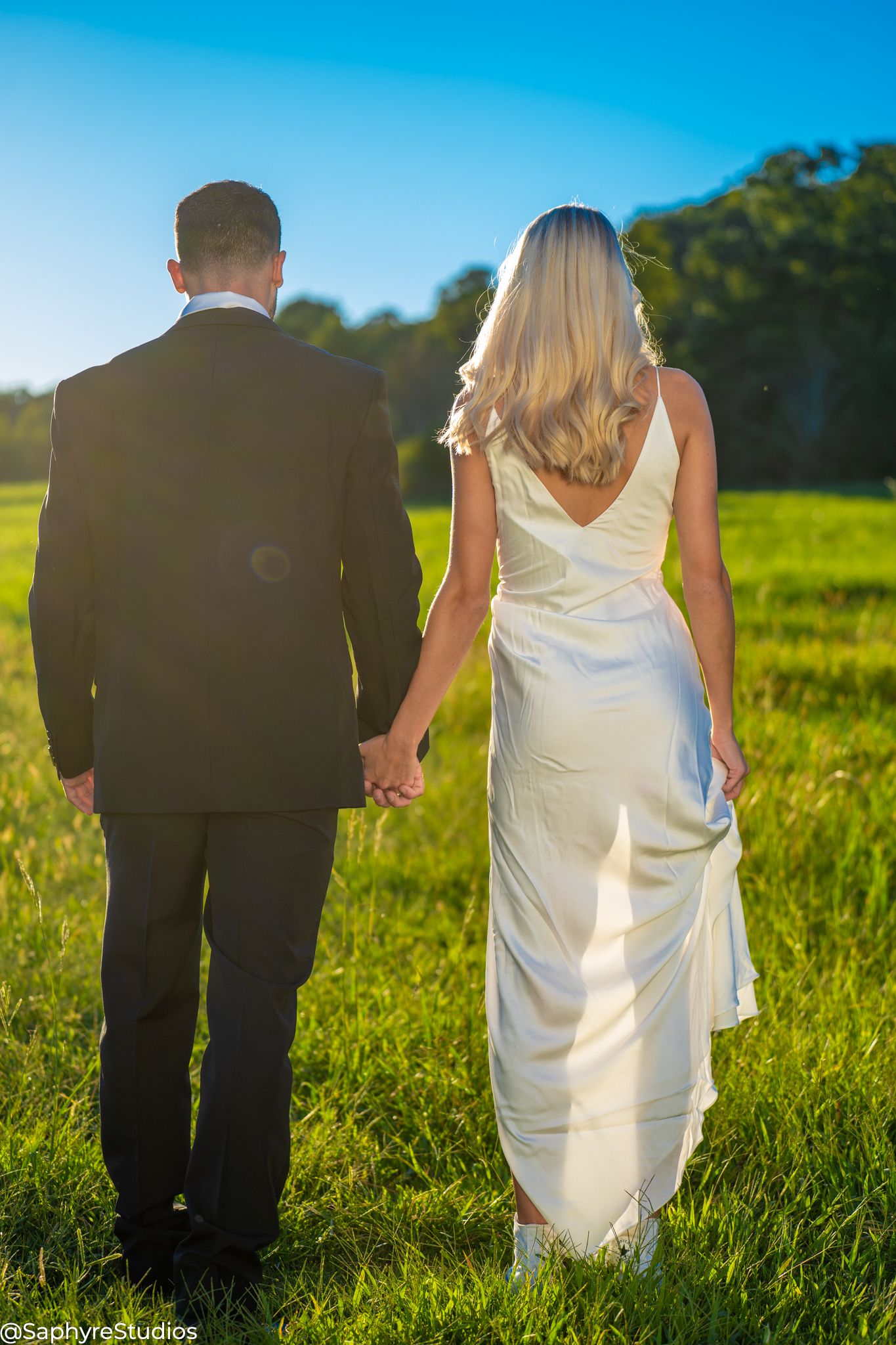 Bride and groom holding hands walking away through sunny green field on their wedding day.