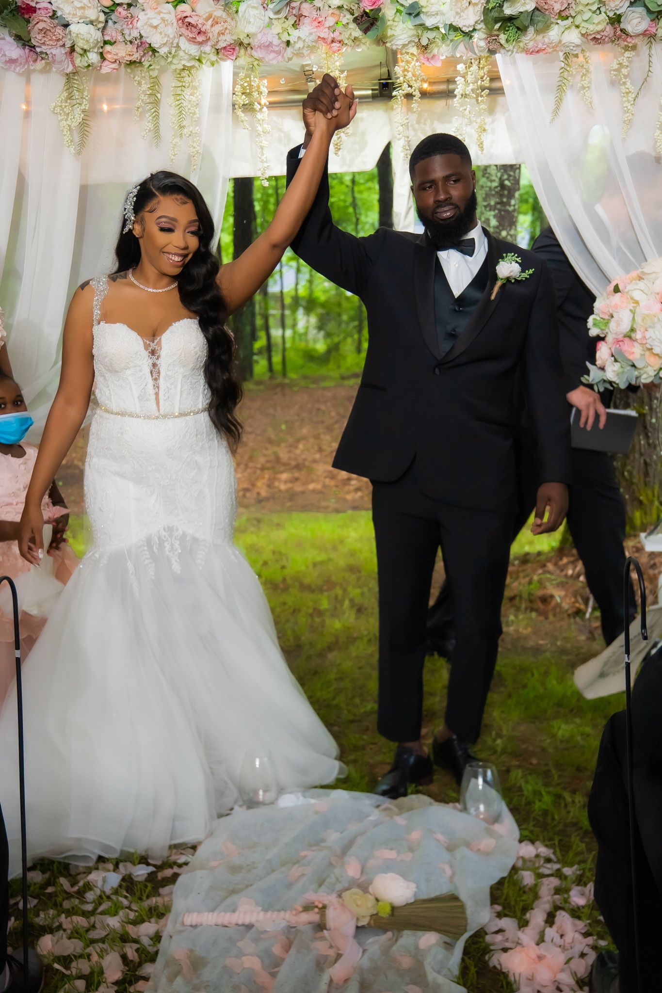 Joyful newlyweds raise hands in celebration under floral arch at outdoor woodland wedding ceremony.