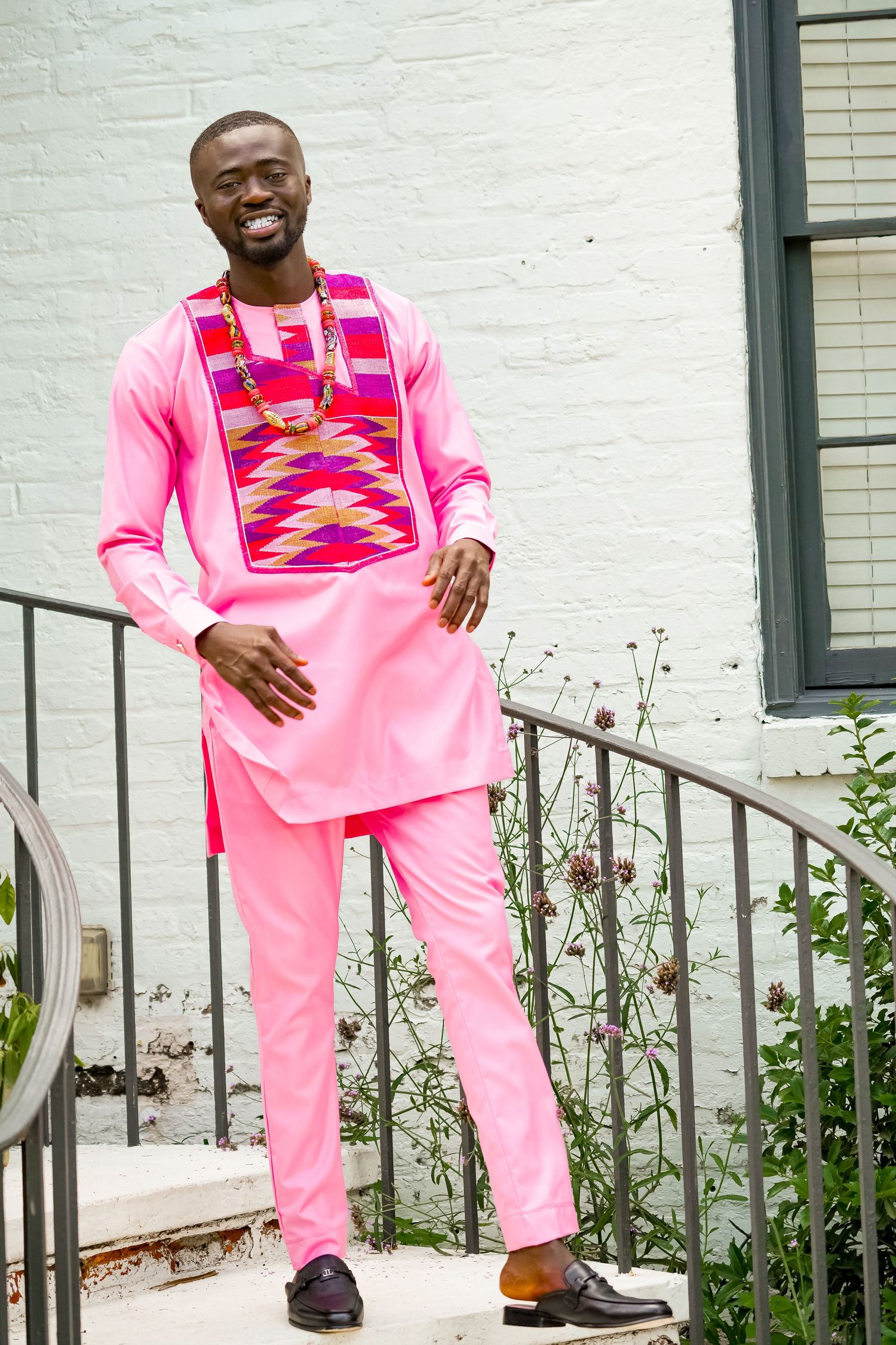 Smiling man in traditional African pink attire with colorful kente embroidery posing on outdoor steps.