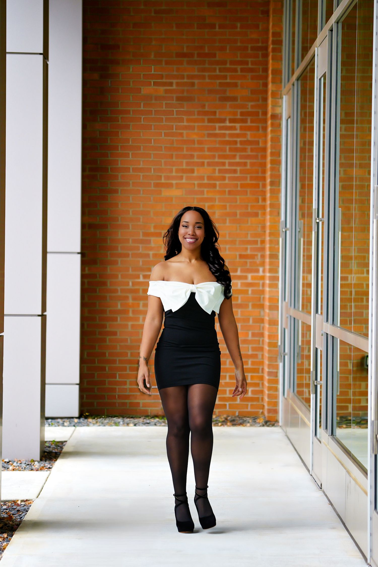Smiling Black woman in black mini dress and white bow top walking near brick wall.