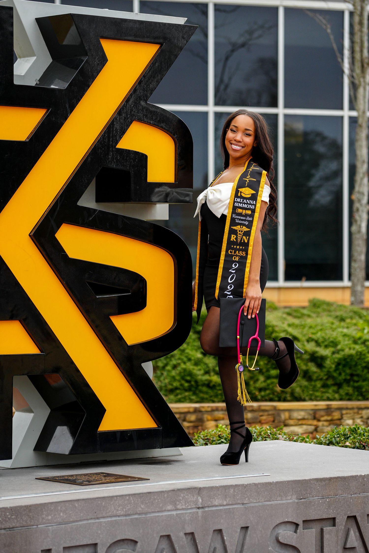 Proud nursing graduate poses beside large KSU logo sign at Kennesaw State University wearing graduation regalia.