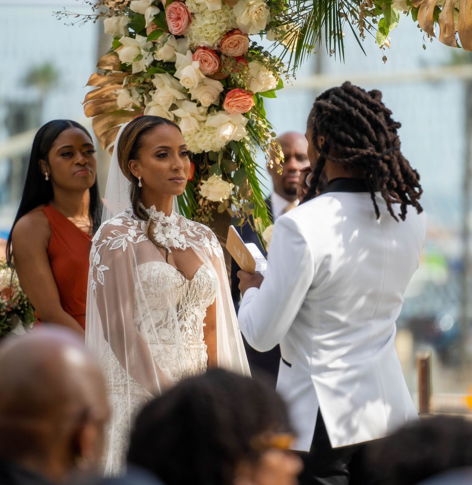Bride in lace gown with cape veil stands at altar with groom in white suit during ceremony.