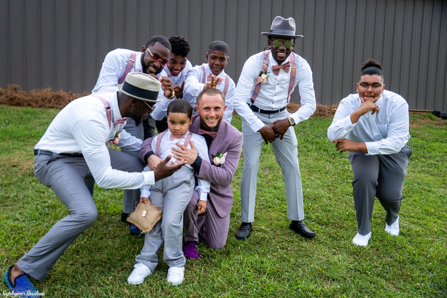 Groom in purple suit laughs with groomsmen in gray suits while posing with young ring bearer outdoors.