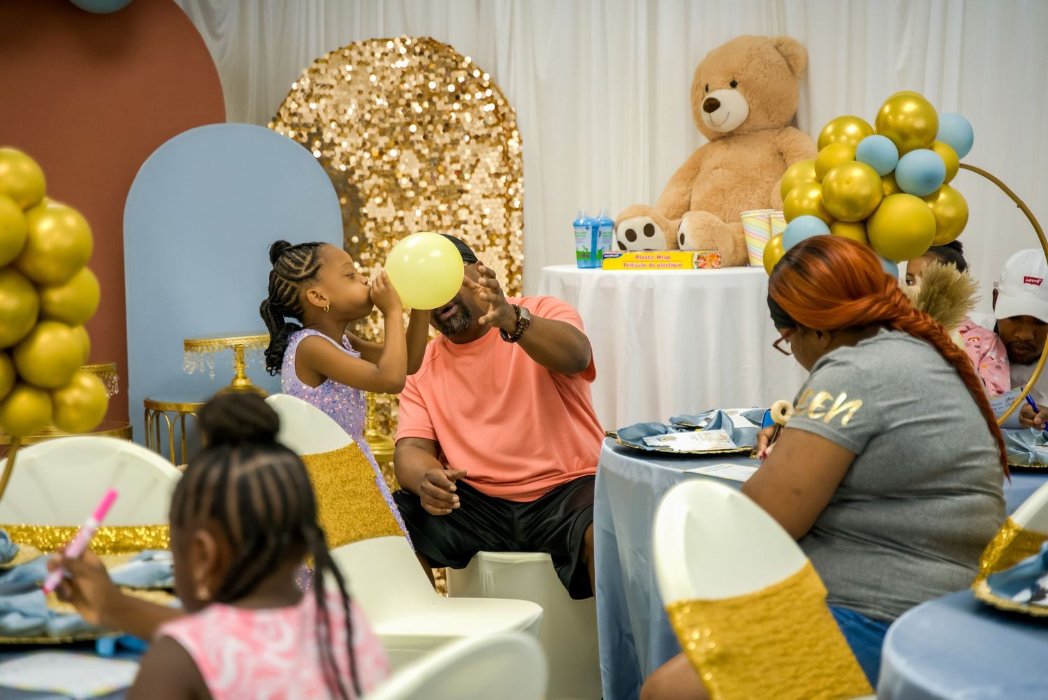 Family enjoying a baby shower with gold and blue balloon decorations and large teddy bear.