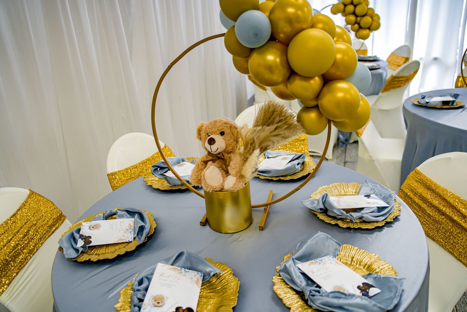 Gold and blue baby shower table with teddy bear centerpiece, balloon arch, and elegant place settings.