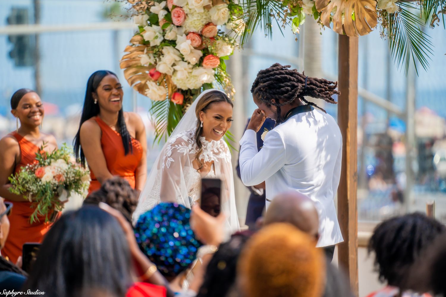 Emotional groom in white suit reacts to brides vows under floral arch at outdoor wedding.