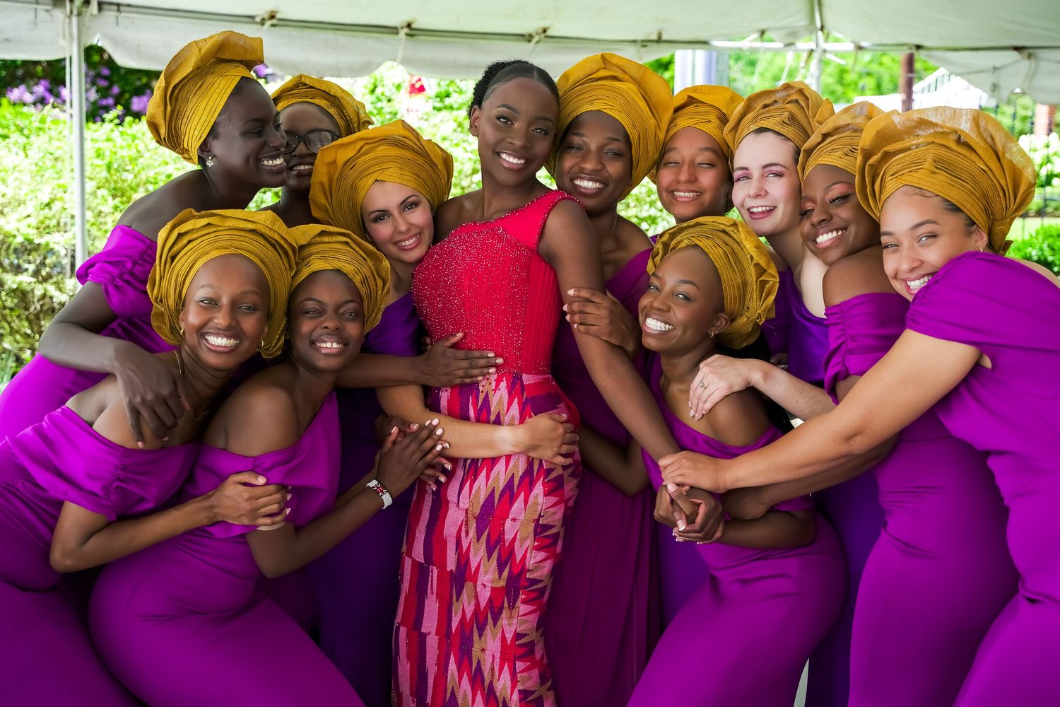 Joyful wedding party in vibrant purple dresses and gold gele headwraps celebrating with bride in red.
