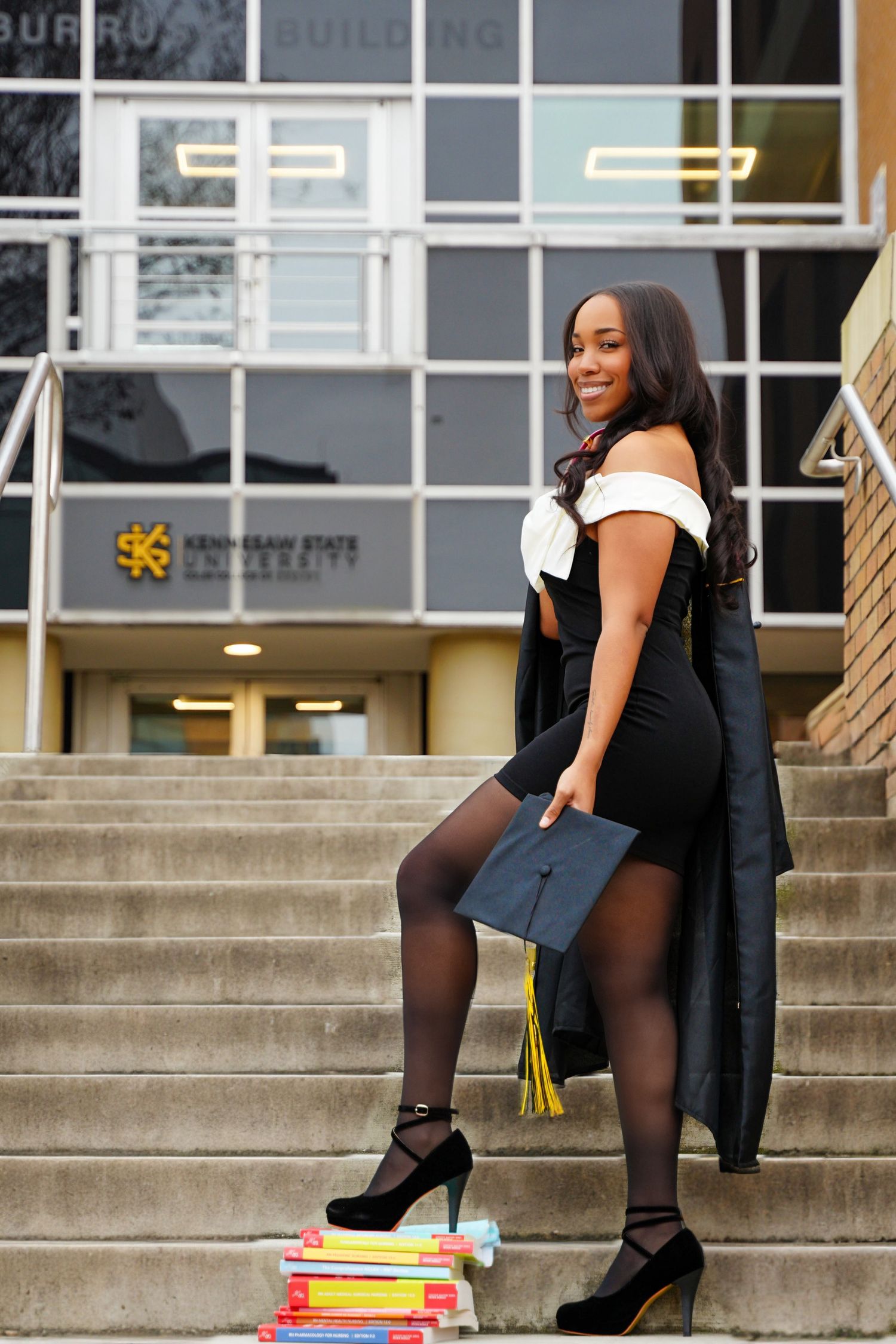 Elegant graduate in black dress holds cap on steps outside Kennesaw State University.