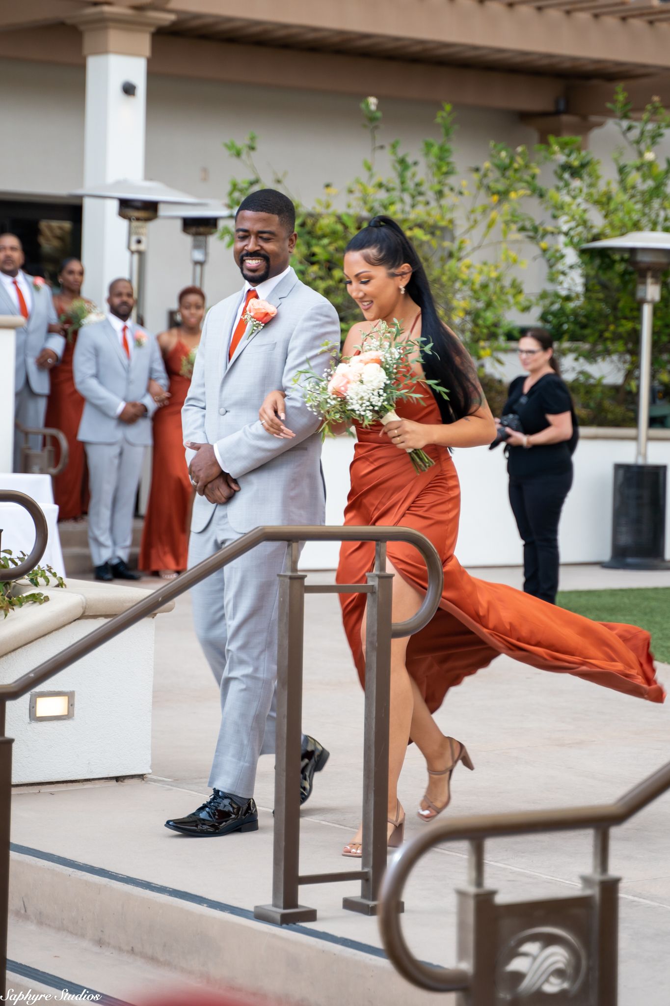 Smiling groomsman in gray suit escorts laughing bridesmaid in orange dress at outdoor wedding ceremony.