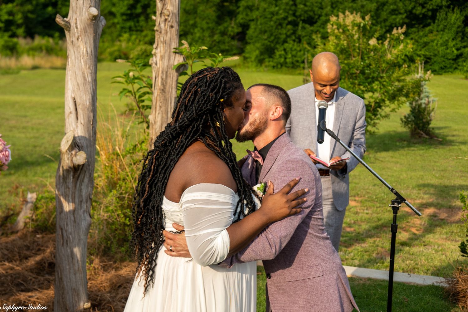 Newlywed couple shares first kiss at outdoor wedding ceremony while officiant watches from behind the altar.