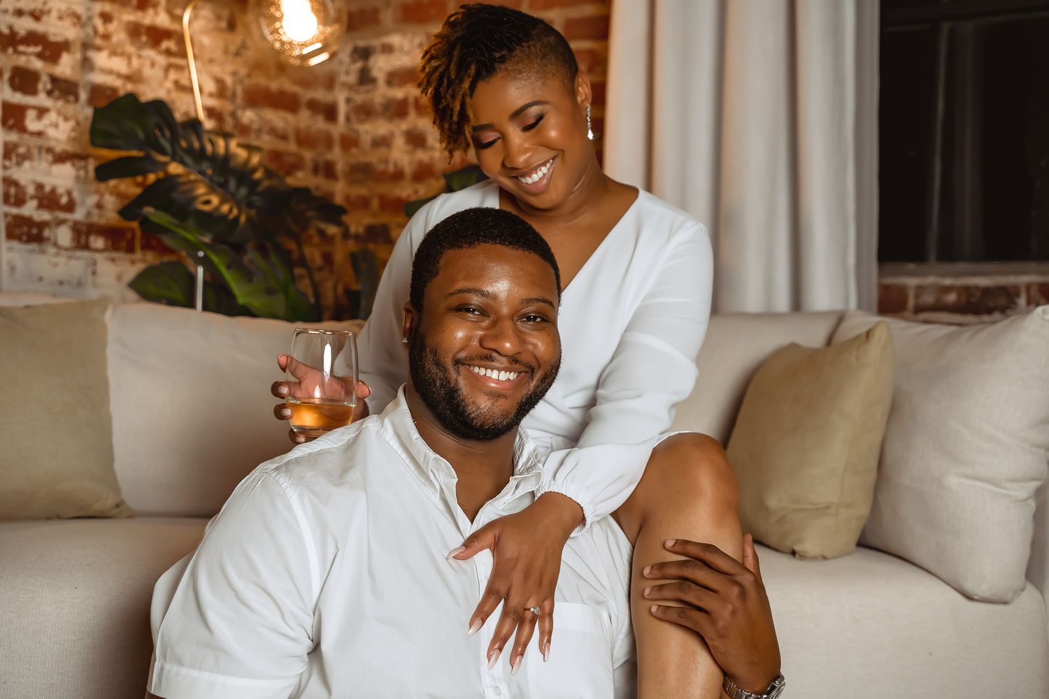 Smiling couple relaxes on living room floor, man holds drink while woman leans over his shoulder.