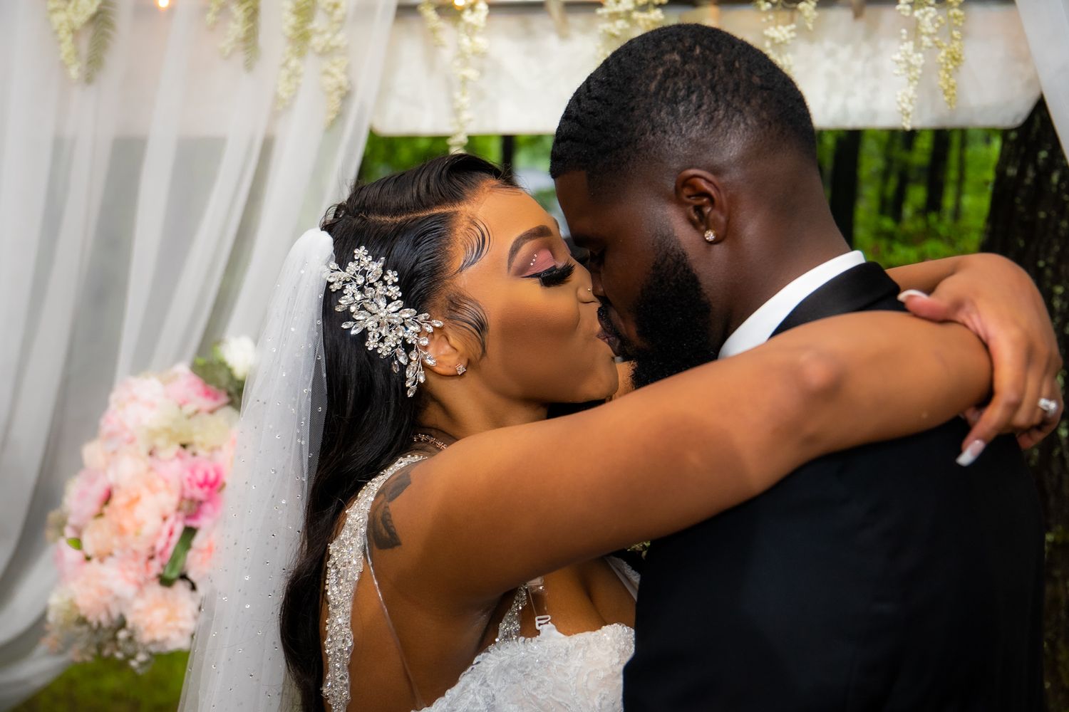 Newlywed couple shares first kiss under floral arch, bride holding pink bouquet at ceremony.