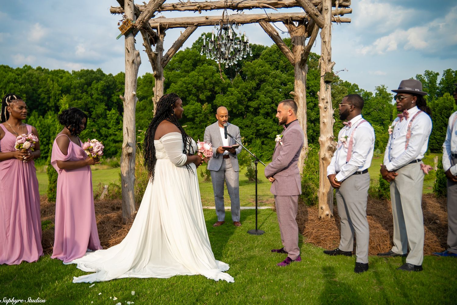 Bride and groom exchange vows under rustic log arch with bridesmaids in pink and groomsmen at outdoor ceremony.
