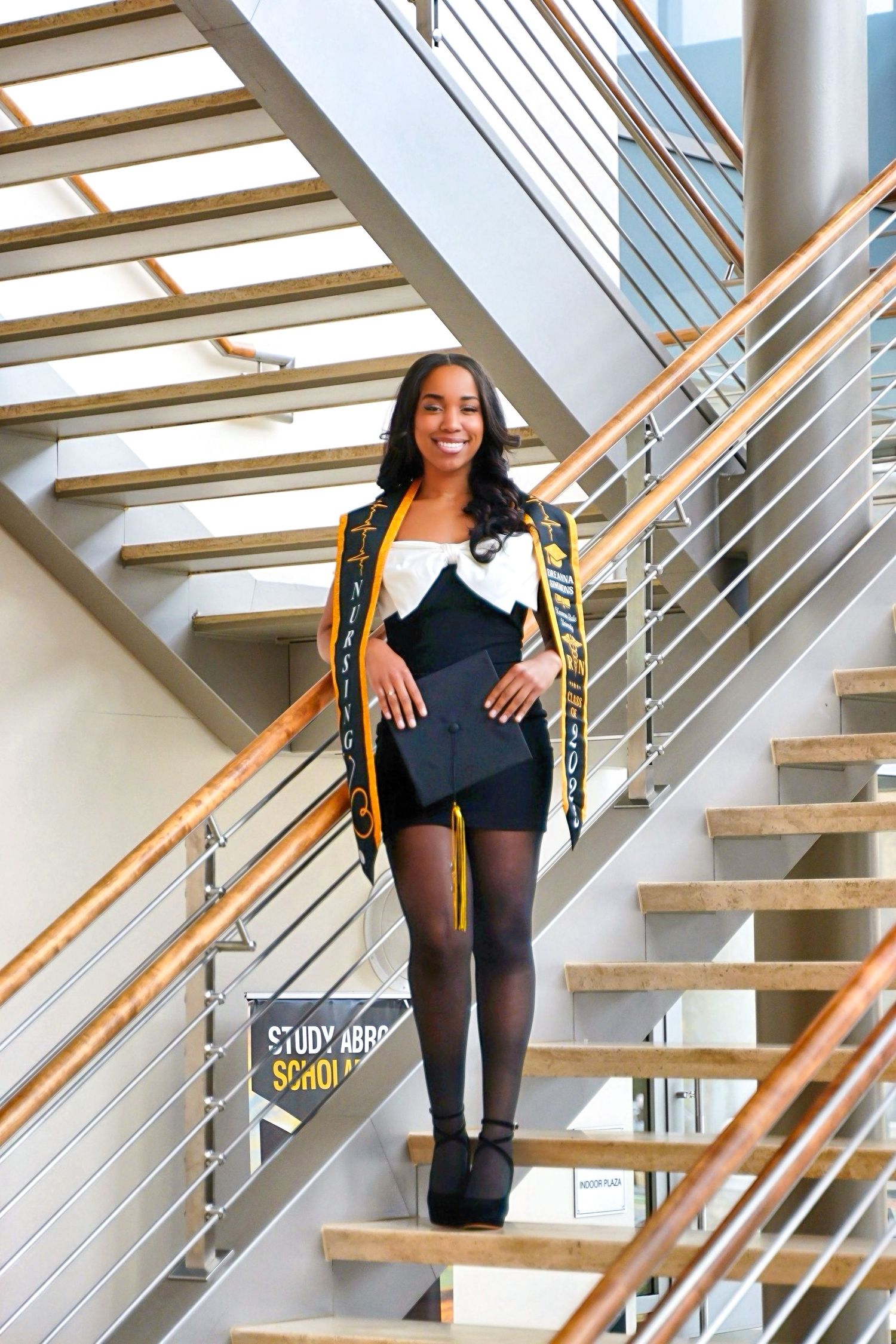 Smiling graduate in black and gold regalia posing on modern staircase holding diploma.
