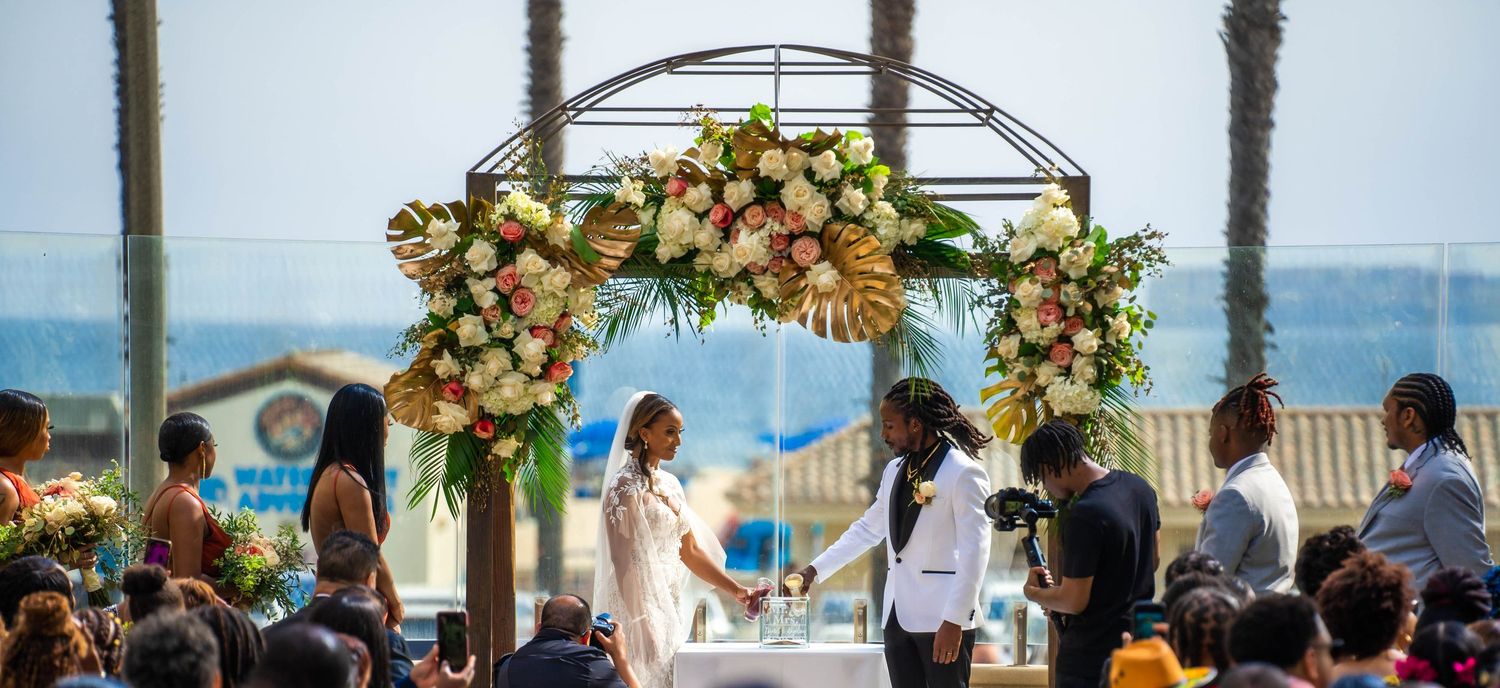 Outdoor beach wedding ceremony under floral arch with couple exchanging vows before seated guests.