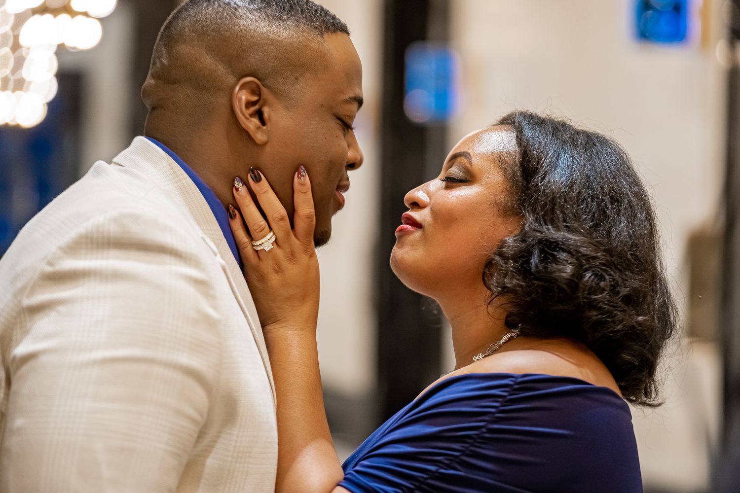 Close romantic moment between couple, woman in blue dress tenderly holds man's face near chandelier.