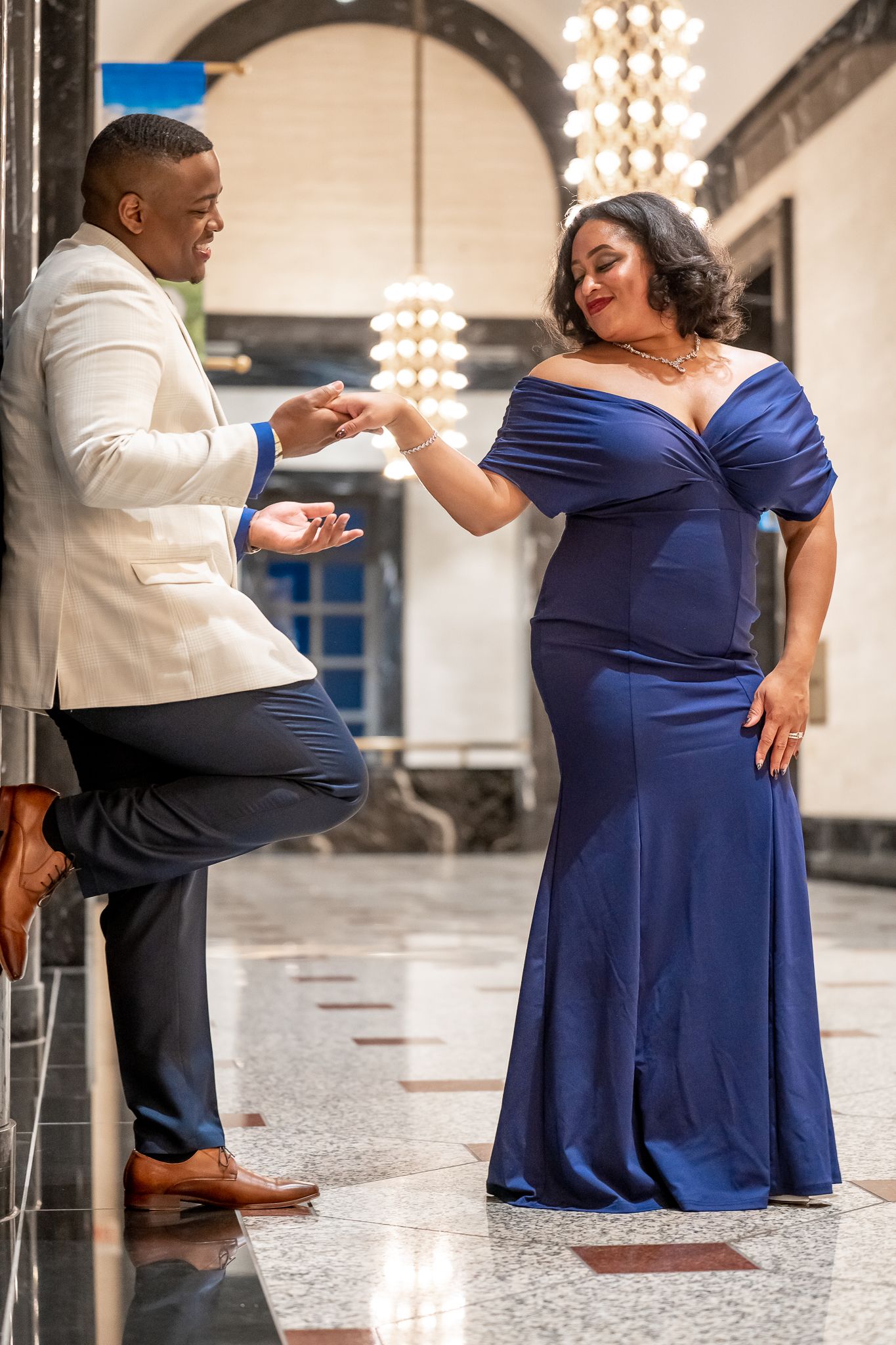 Elegant couple poses in grand hall, man in cream blazer holds woman's hand as she wears blue gown.