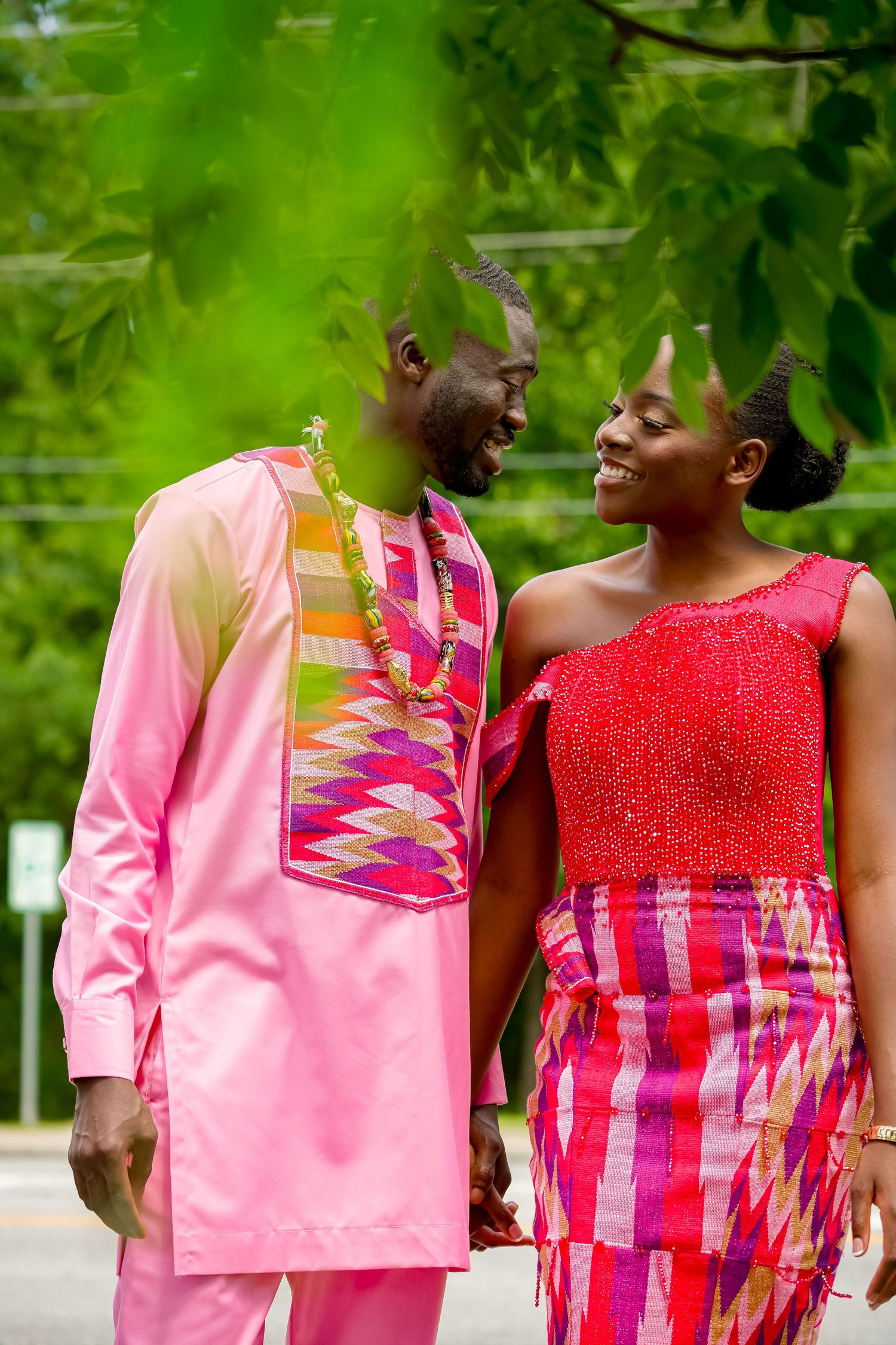 A smiling couple in pink and red African traditional outfits walk together outdoors.