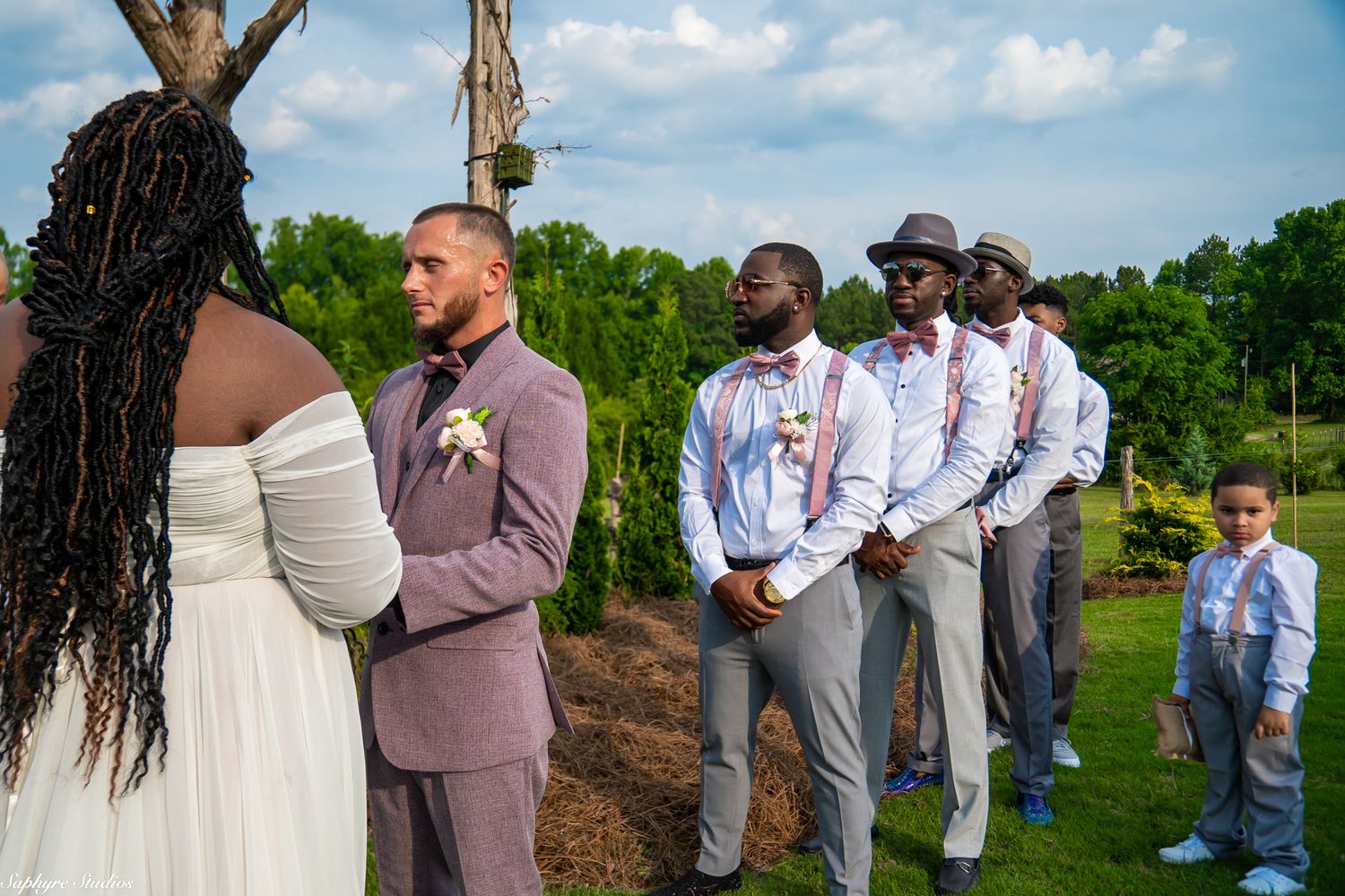 Groom in mauve suit stands at altar with groomsmen in gray suits and young ring bearer during outdoor wedding.