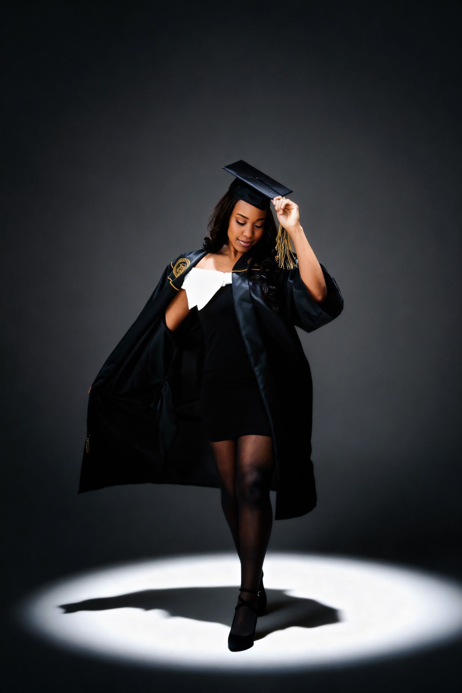 African American woman in graduation cap and gown posing dramatically in spotlight on dark background.