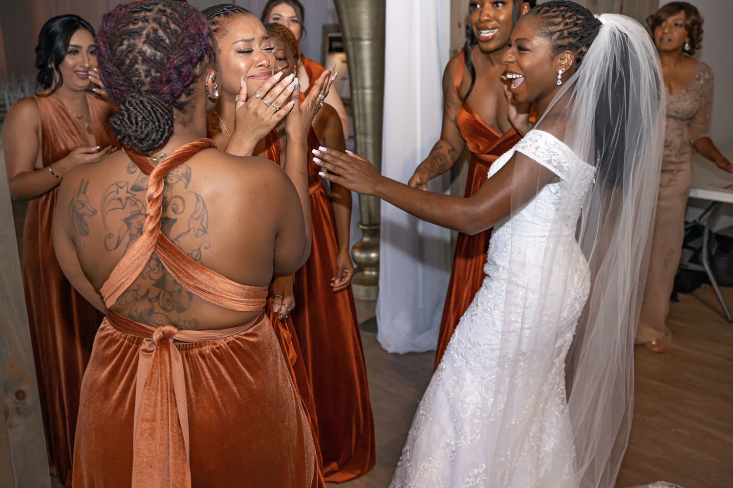 Bride in white lace wedding gown sharing emotional moment with bridesmaid in rust velvet dress indoors.