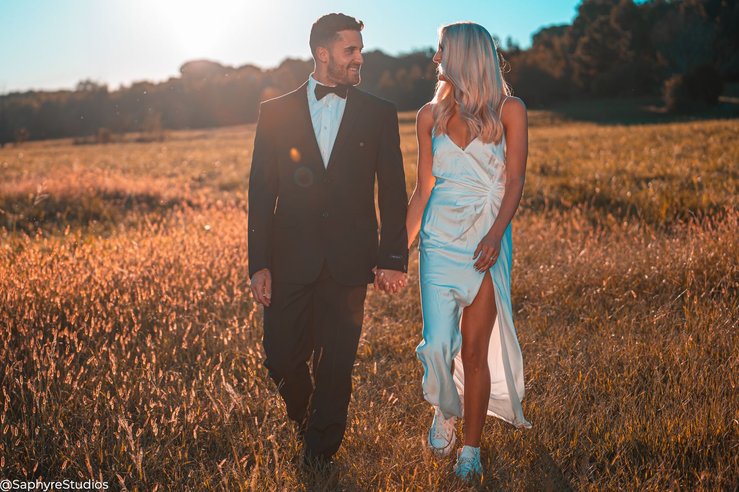 Bride and groom holding hands walking through golden field at sunset in elegant wedding attire.