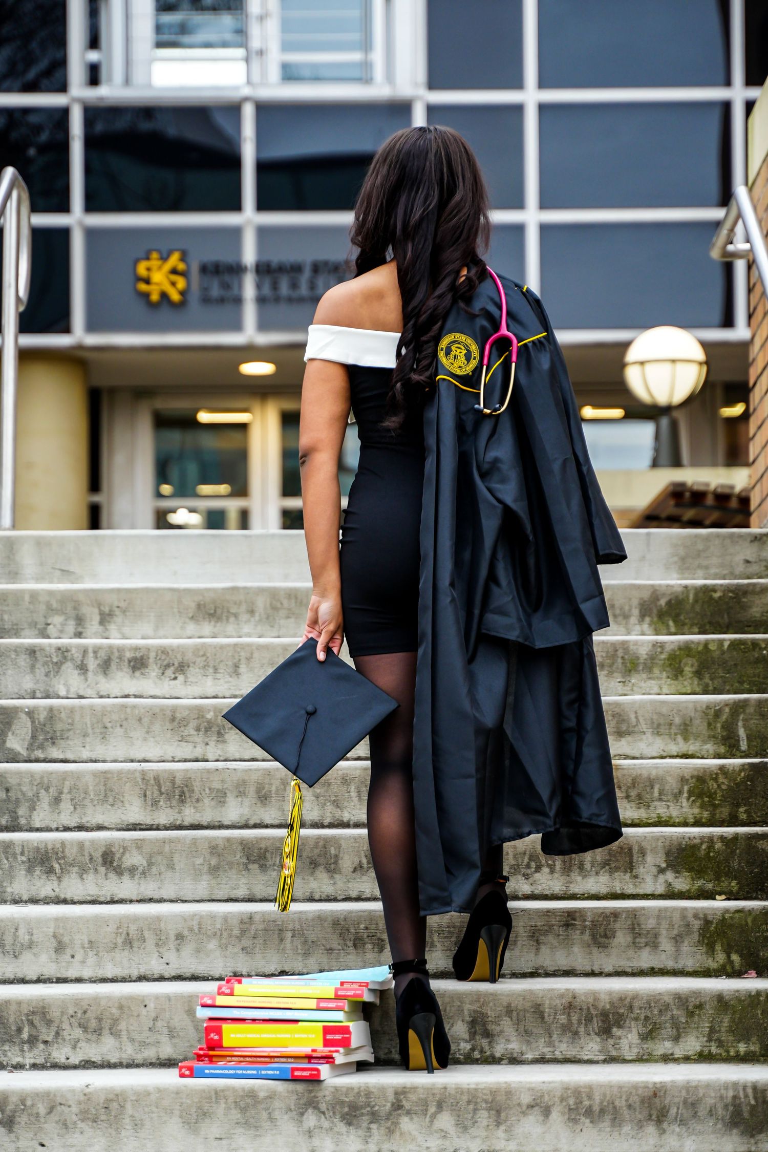 Nursing graduate poses on steps with cap, gown, stethoscope, and colorful textbooks at Kennesaw State University.