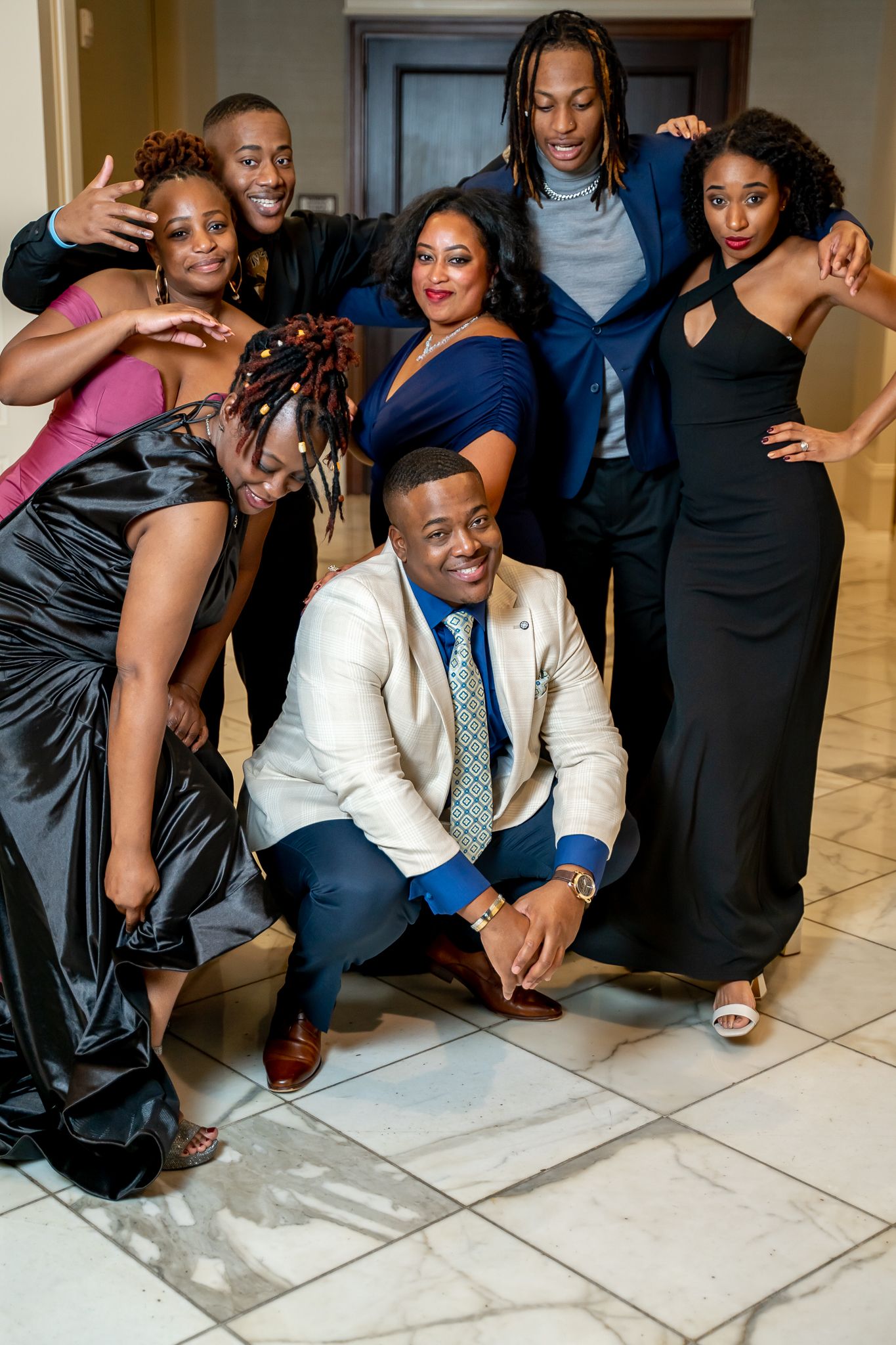 Seven stylish friends in formal wear posing playfully together at an elegant indoor event.