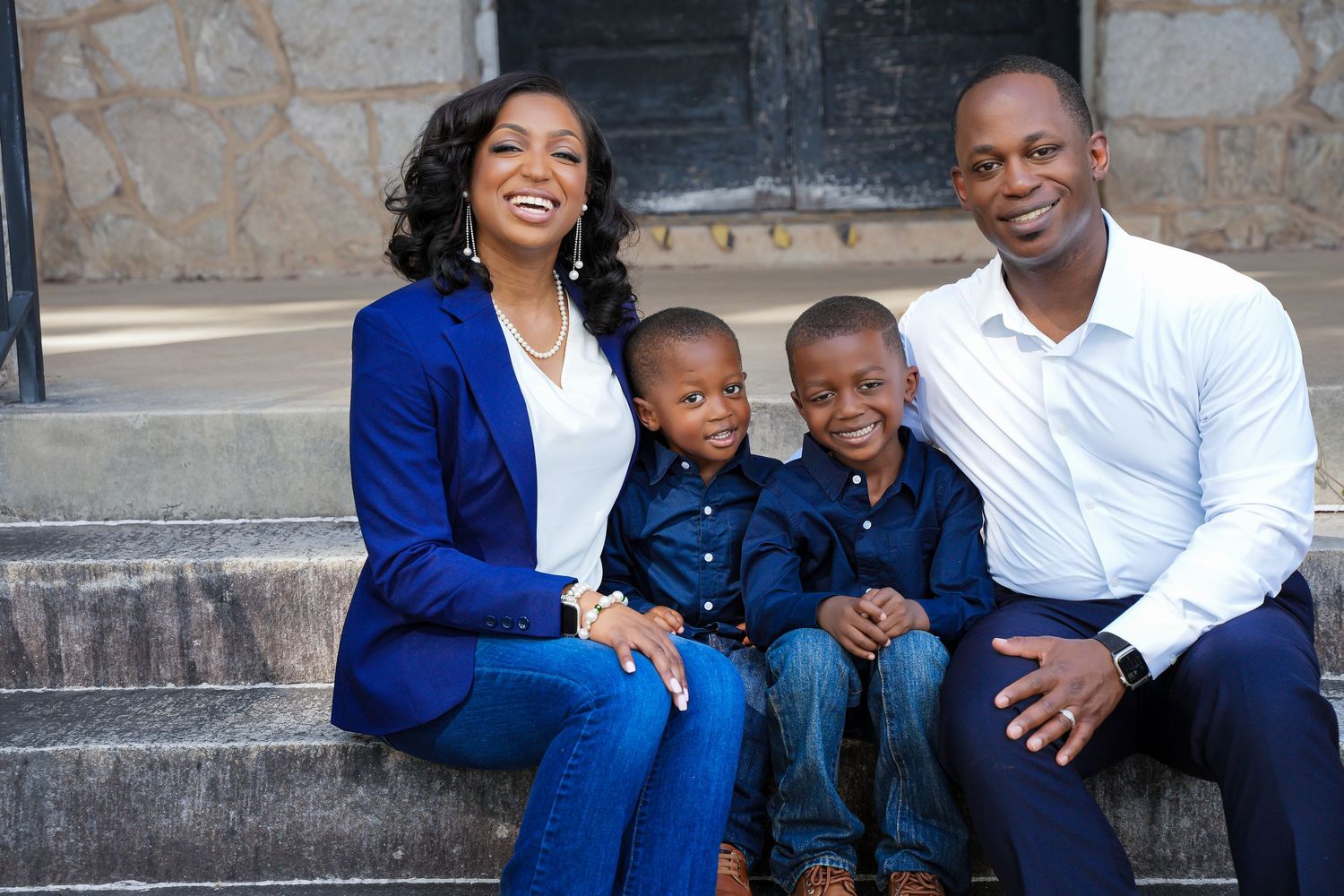 Smiling African American family of four sitting on stone steps wearing coordinated blue outfits outdoors.