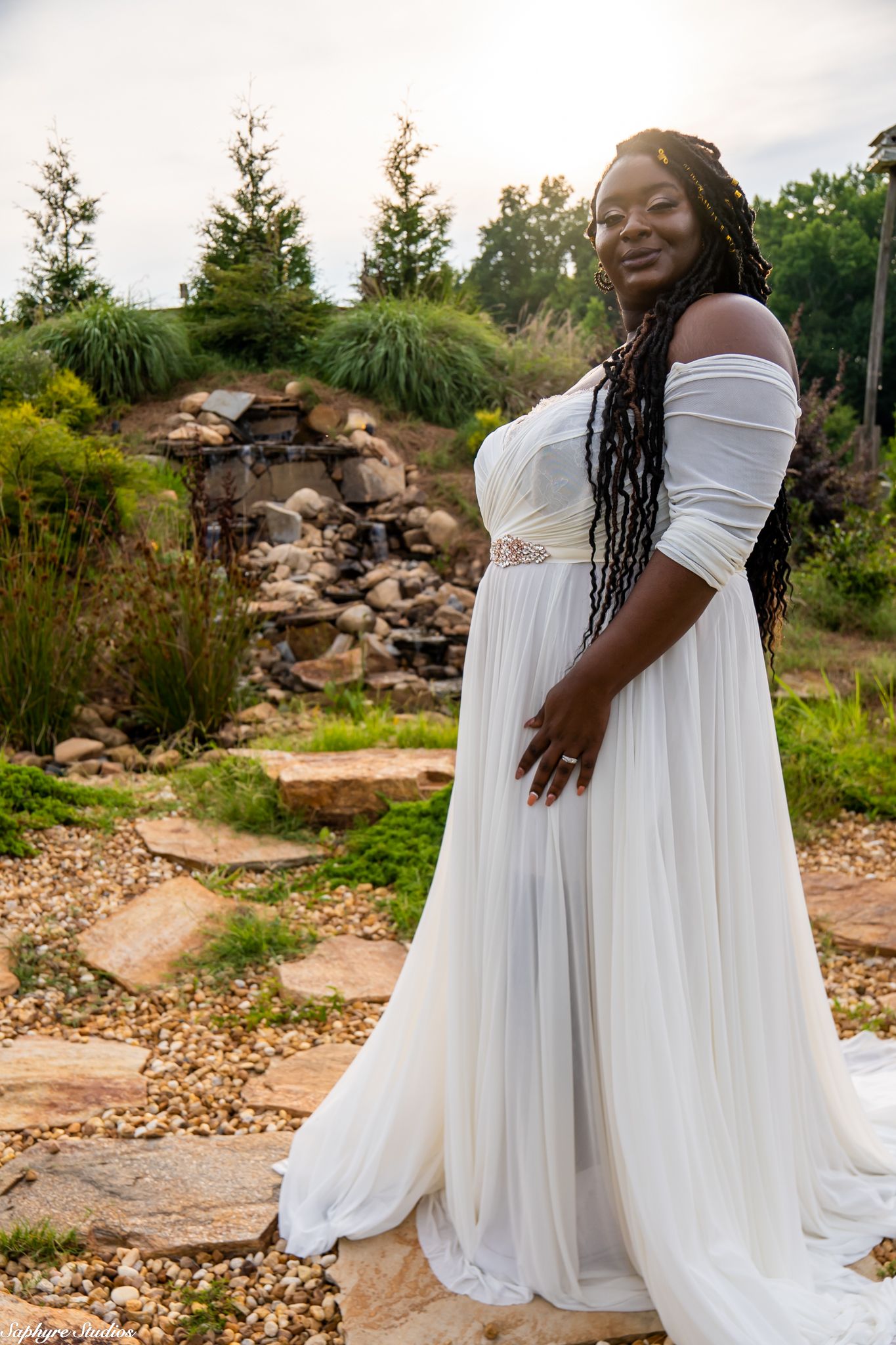 Bride with long braids smiles in elegant white off-shoulder wedding gown posed near stone waterfall garden feature.