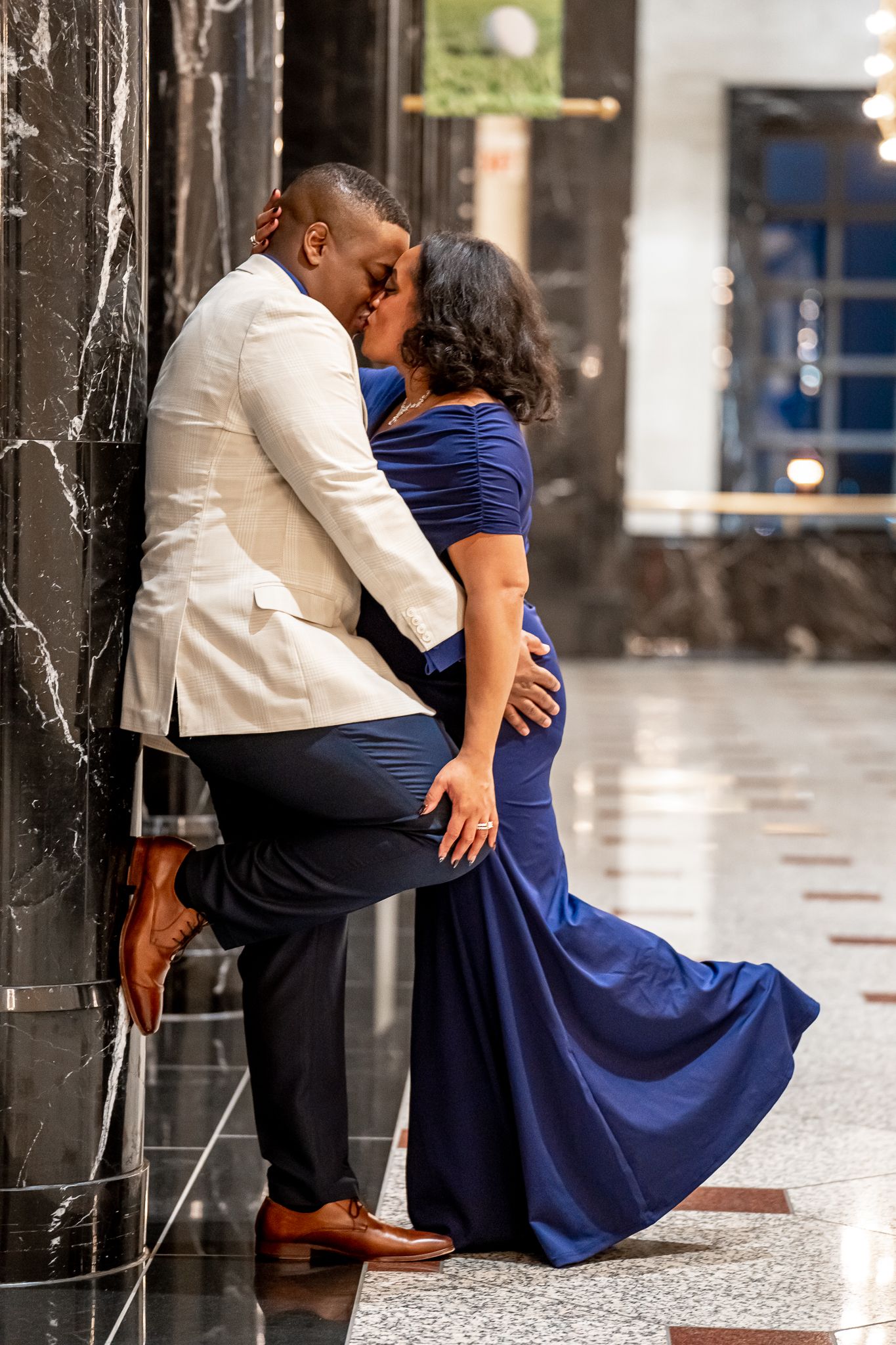 Couple shares romantic kiss against marble wall, woman in blue mermaid gown, man in cream blazer.