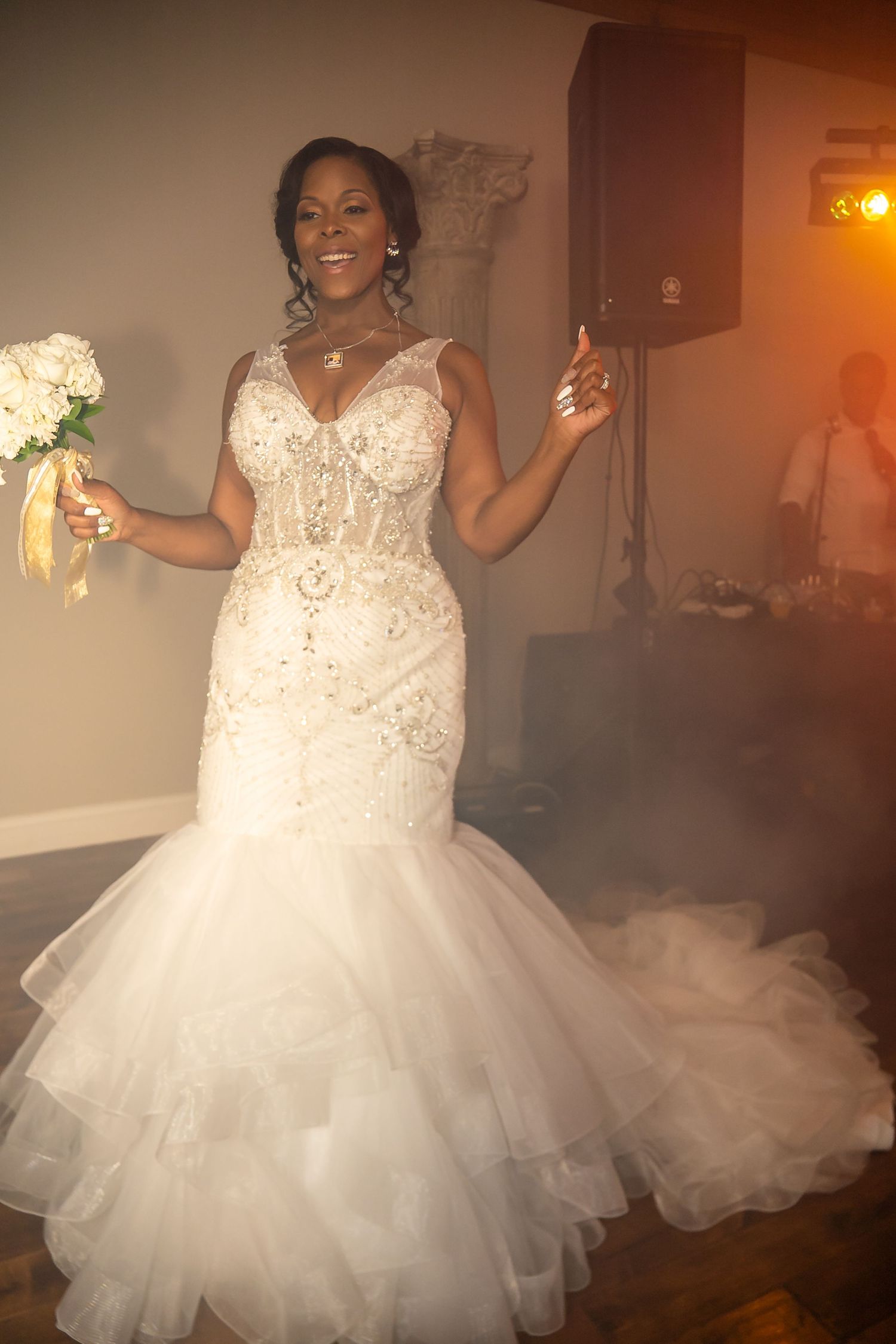 Radiant bride in beaded mermaid gown holds bouquet on smoky dance floor.