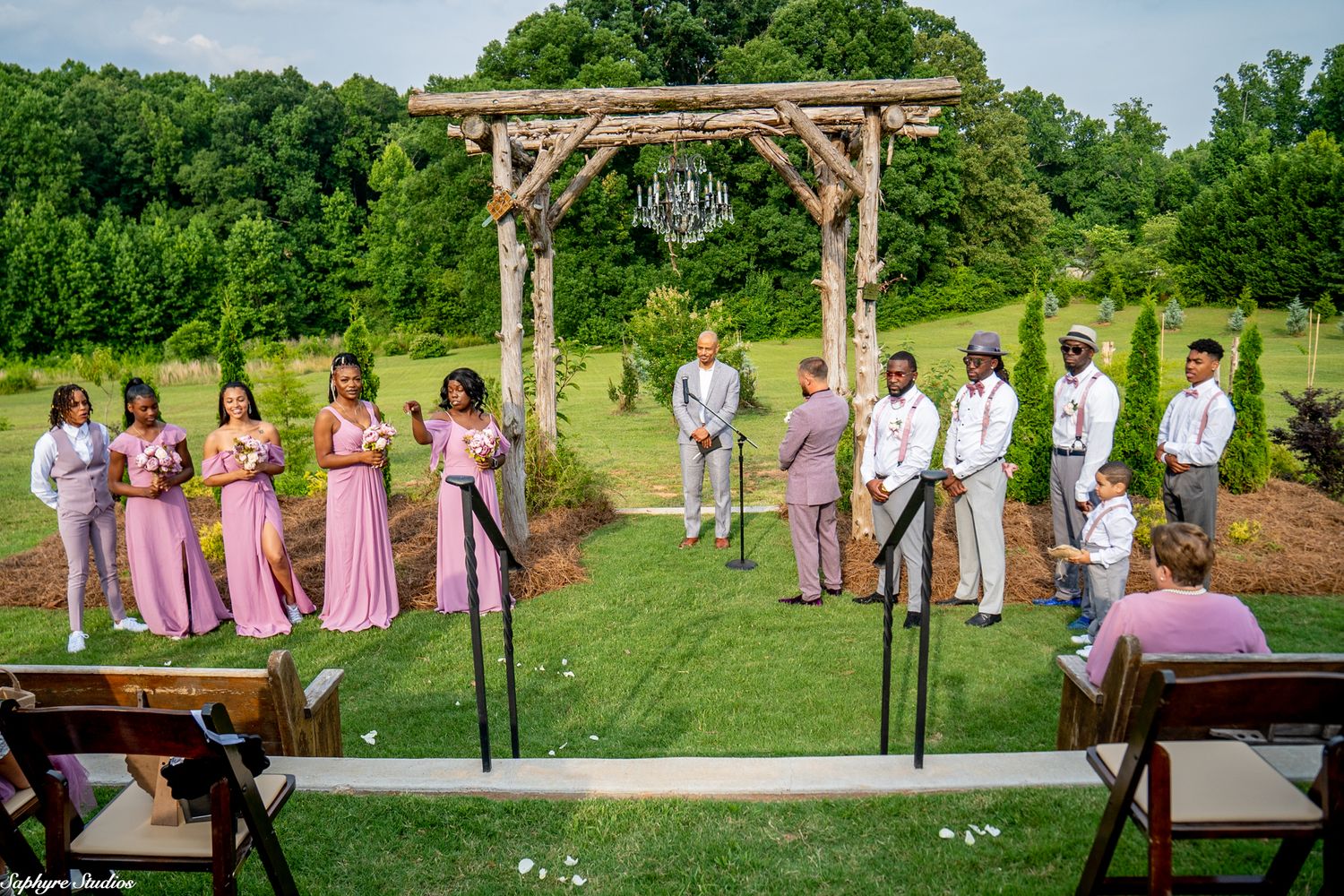 Wedding party stands at rustic wooden arch altar on green lawn with bridesmaids in pink and groomsmen in gray.