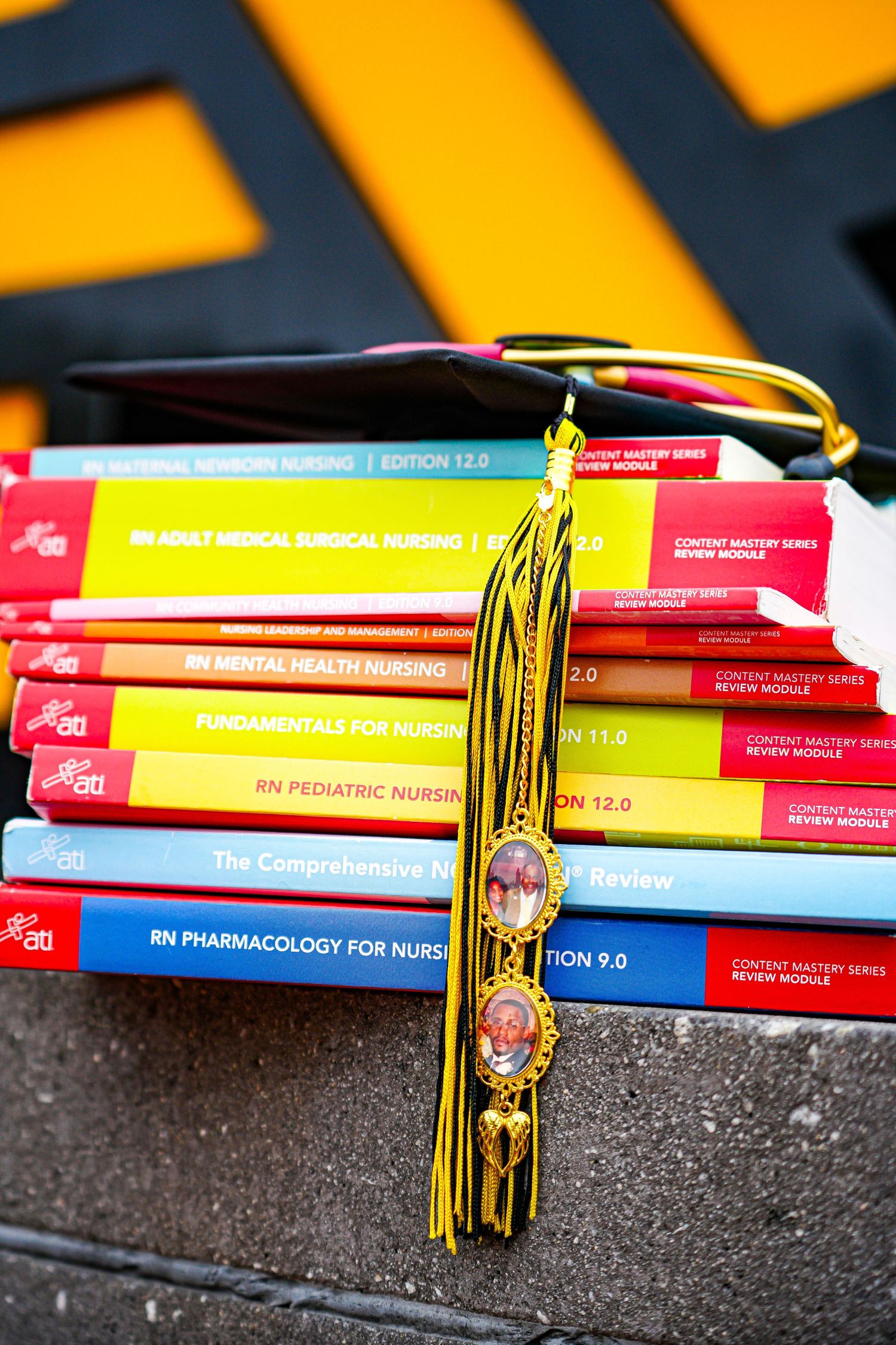 Nursing graduation cap with gold tassel and memorial locket displayed on stack of colorful ATI nursing textbooks.