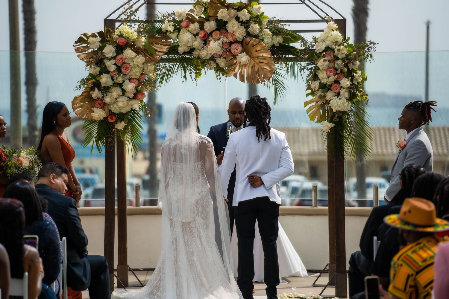 Bride in cathedral veil and groom exchanging vows under elegant floral arch at outdoor beach ceremony.