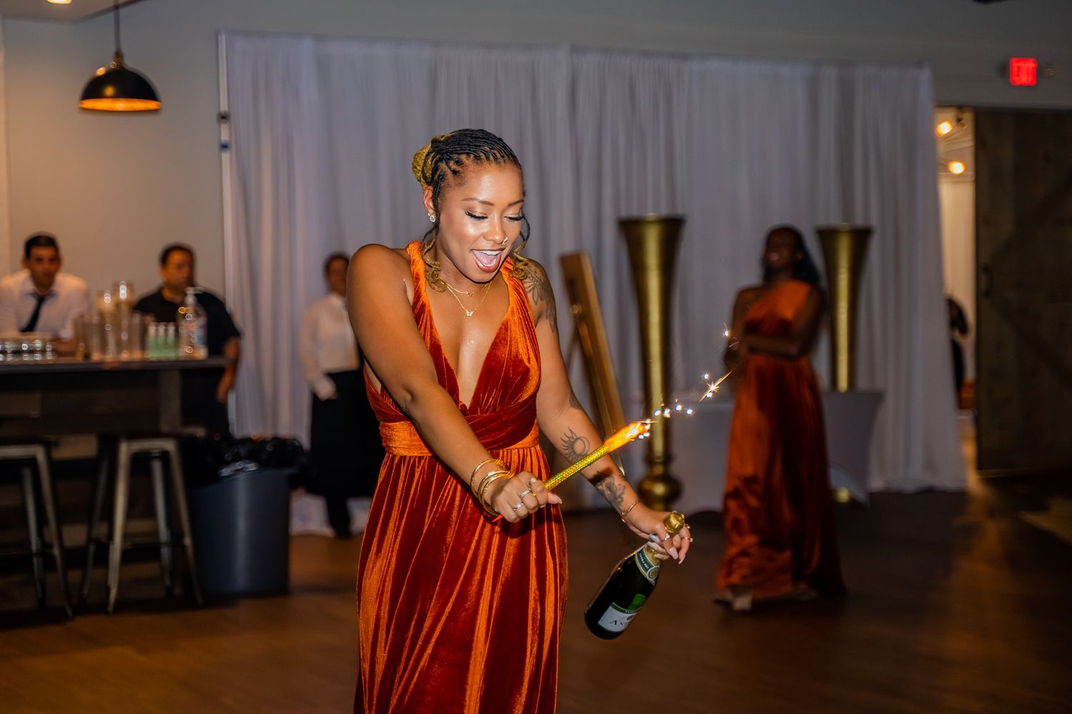 Woman in rust velvet gown sabering champagne bottle at elegant wedding reception celebration.