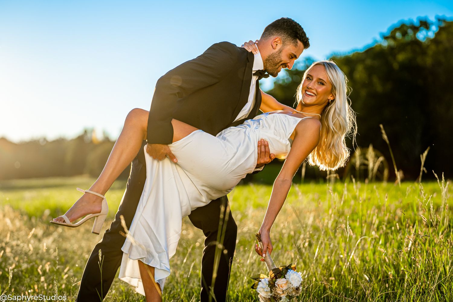 Joyful groom lifts laughing bride in sunny green meadow during outdoor wedding photo session.