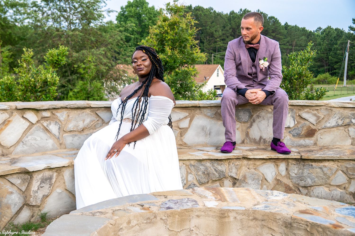 Bride and groom pose on stone wall outdoors, bride in white gown, groom in lavender suit.