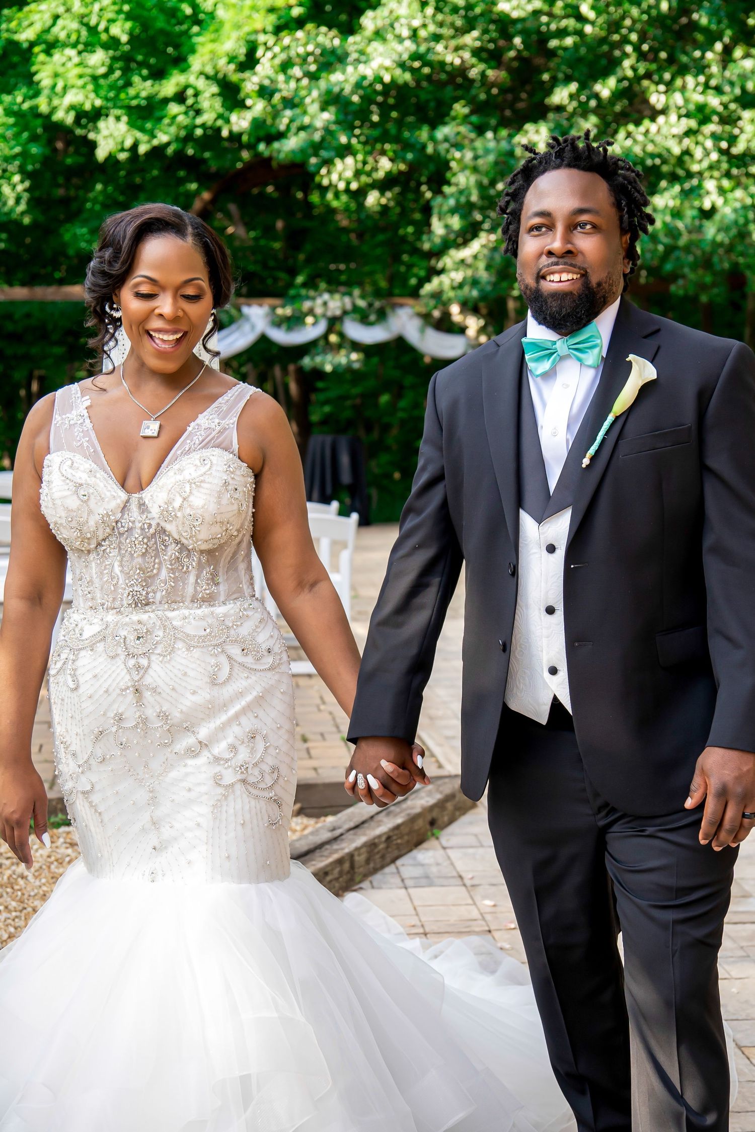 Smiling bride and groom holding hands walking outdoors at wedding ceremony, bride in beaded mermaid gown, groom in black tuxedo.