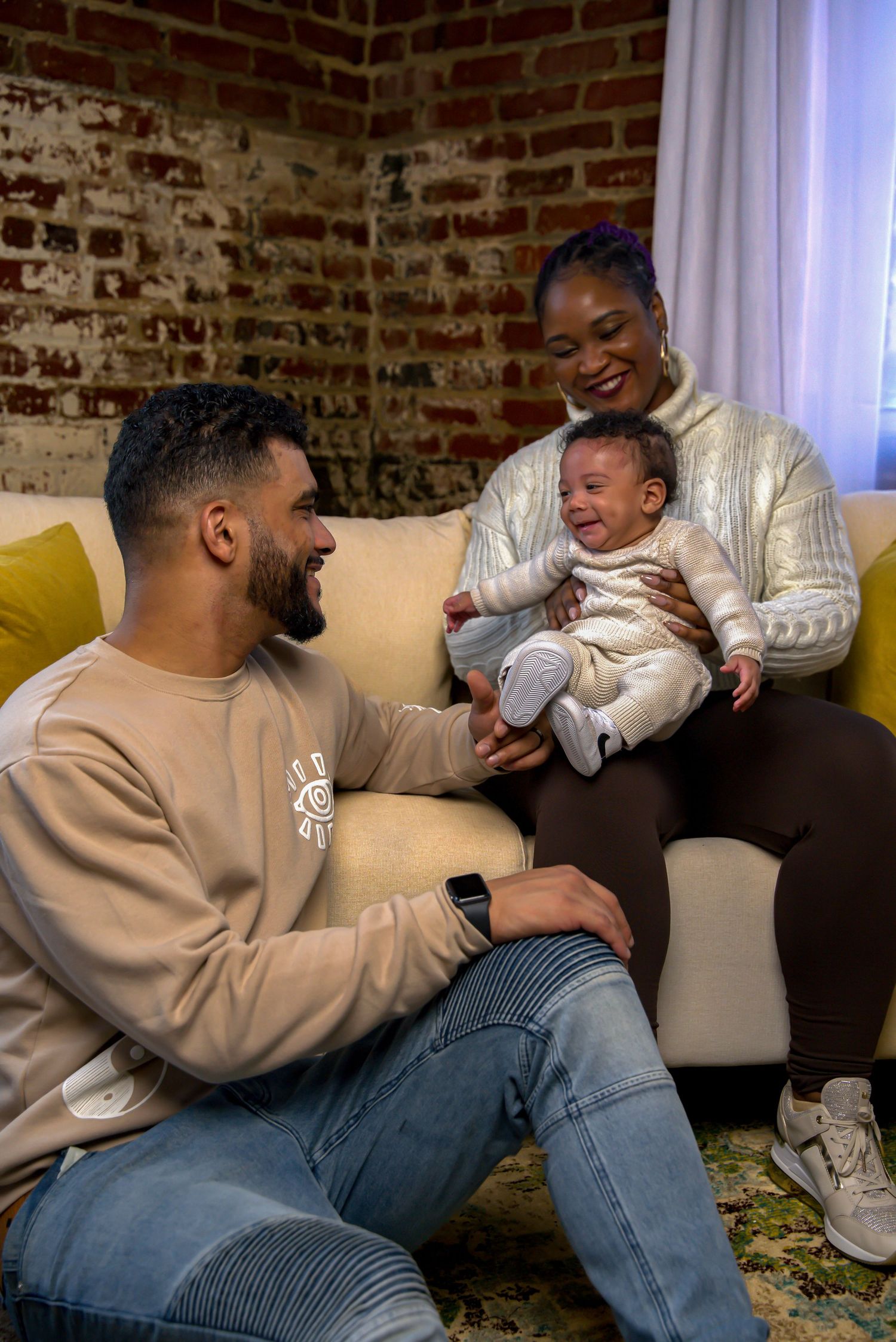 Young family smiling together indoors, parents admiring laughing newborn baby on cozy couch.