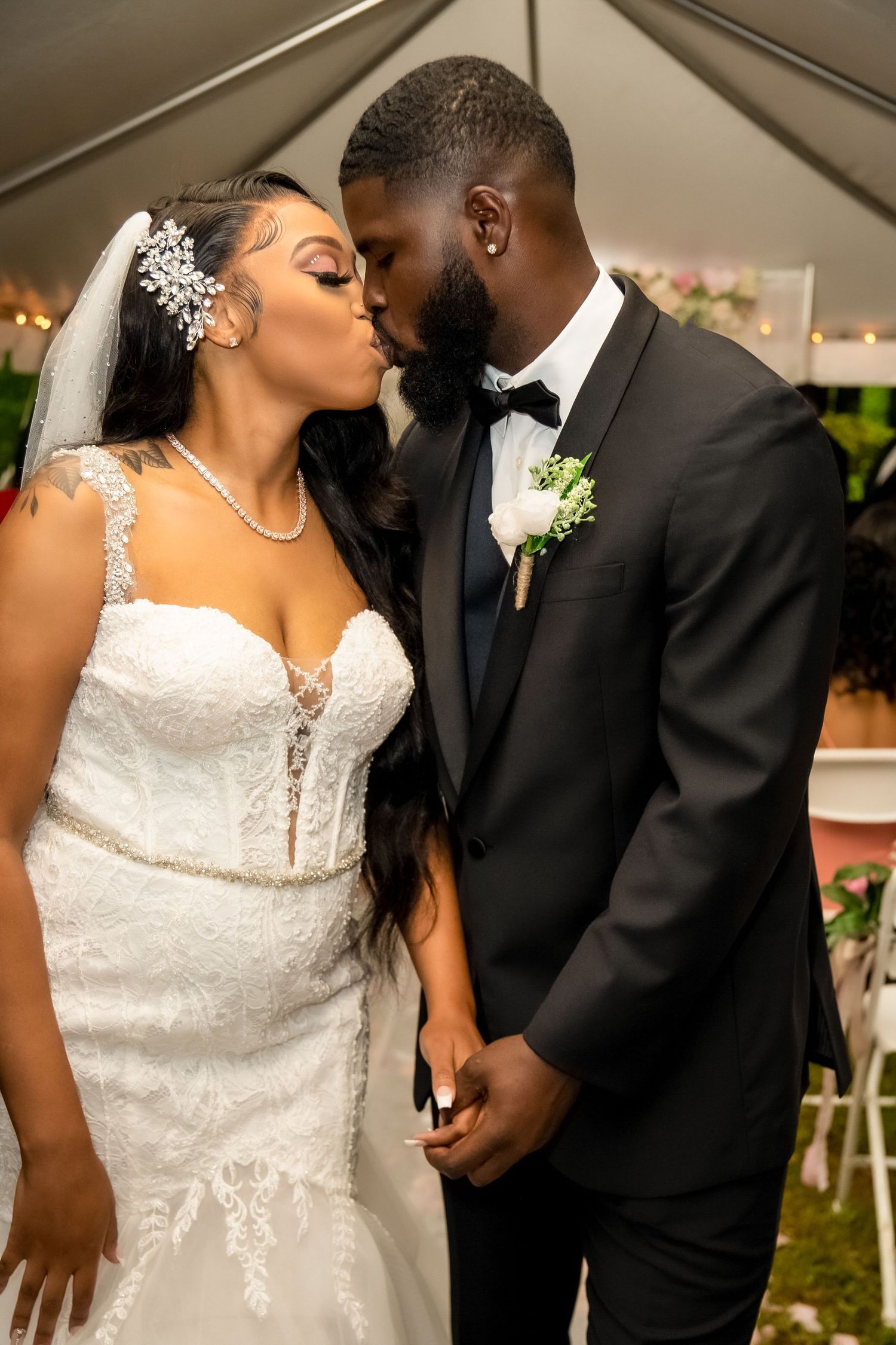 Bride and groom sharing first kiss under tent at wedding reception, bride in lace mermaid dress, groom in black tuxedo.