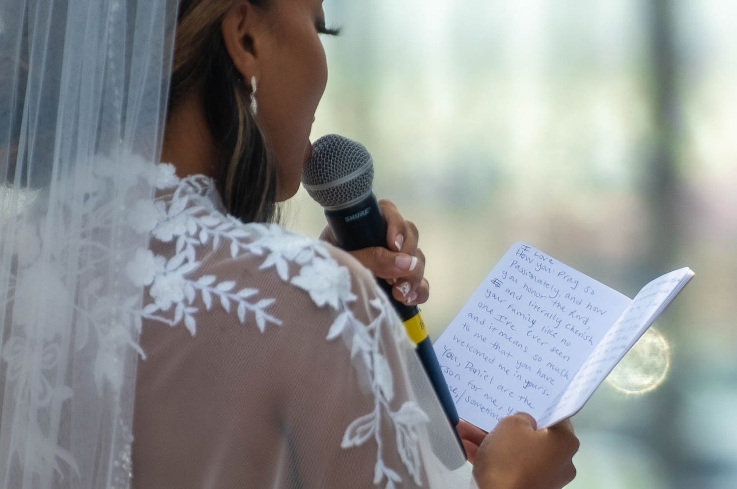 Bride in lace veil reading handwritten wedding vows into microphone during outdoor ceremony.