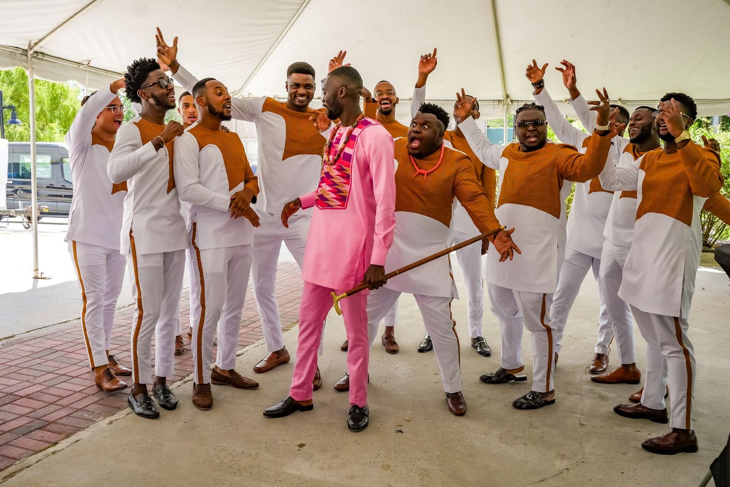 Groom in pink suit celebrates joyfully with groomsmen in white and orange traditional outfits.