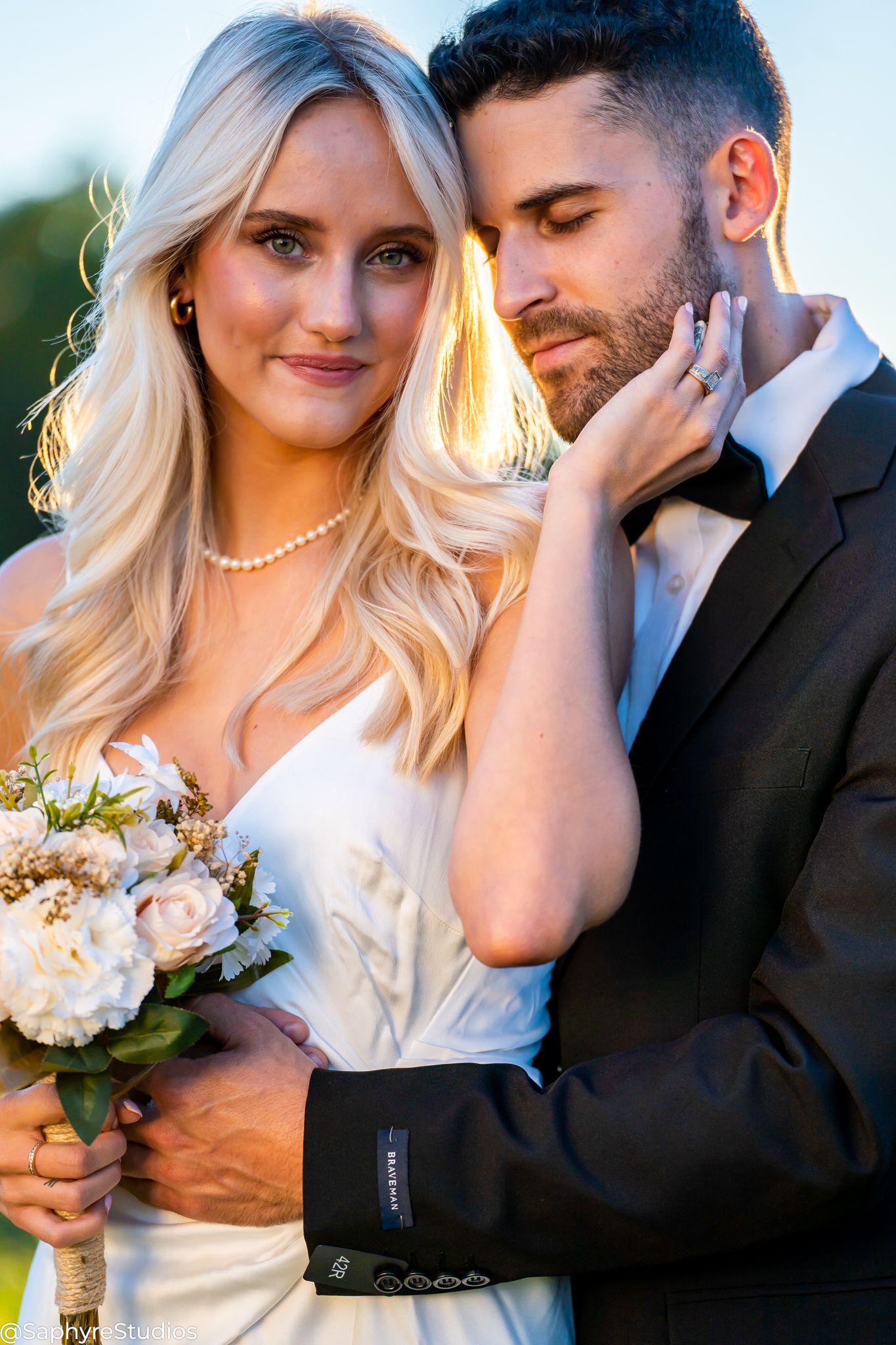 Bride holding white bouquet leans into groom during romantic golden hour wedding portrait.