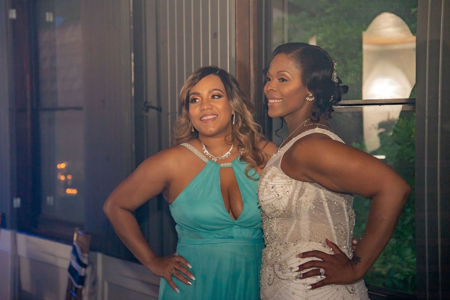 Two women posing together at a wedding, one in teal dress and one in silver beaded gown.