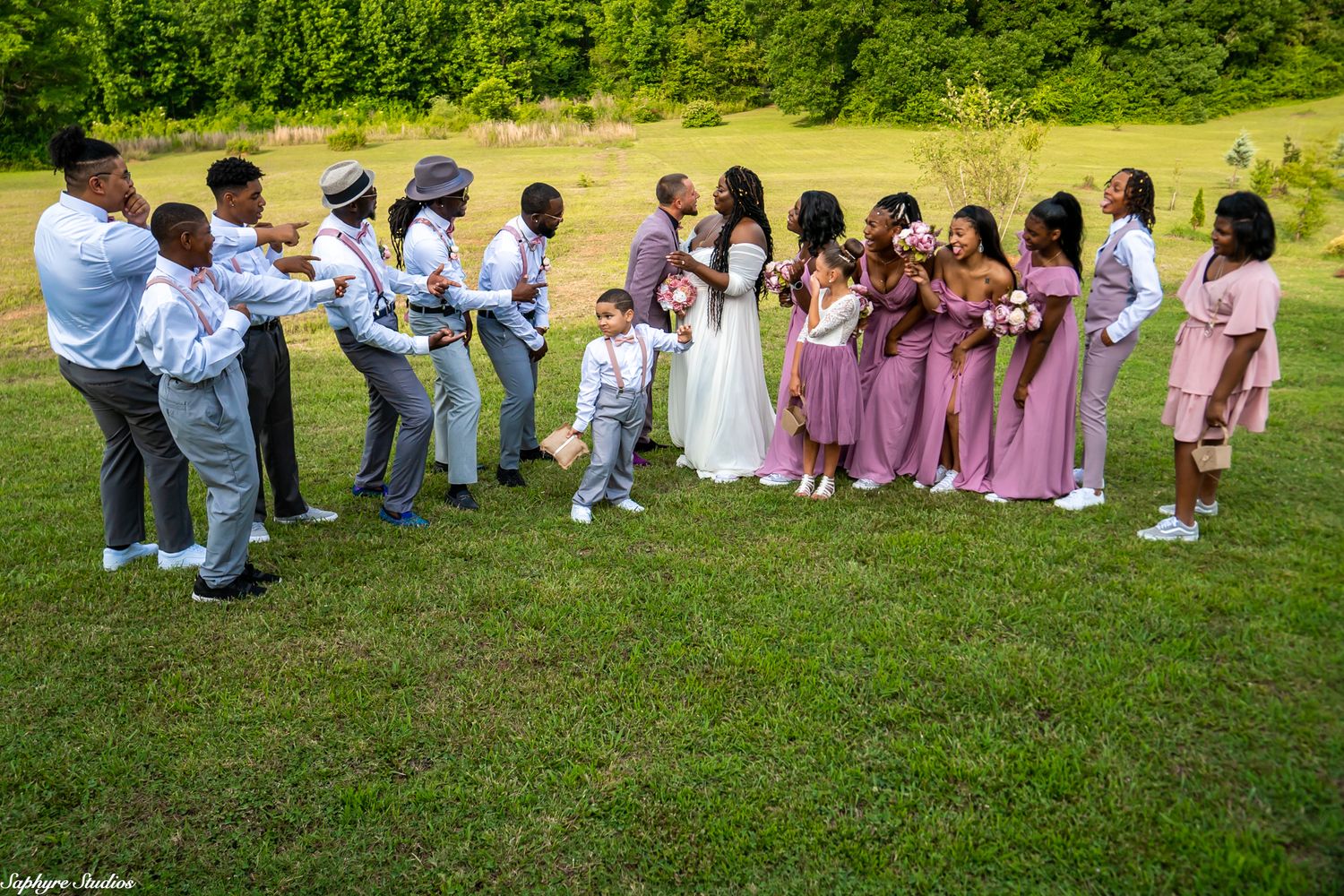 Wedding party faces each other in two lines on grass, groomsmen in gray and bridesmaids in mauve with bride and groom center.